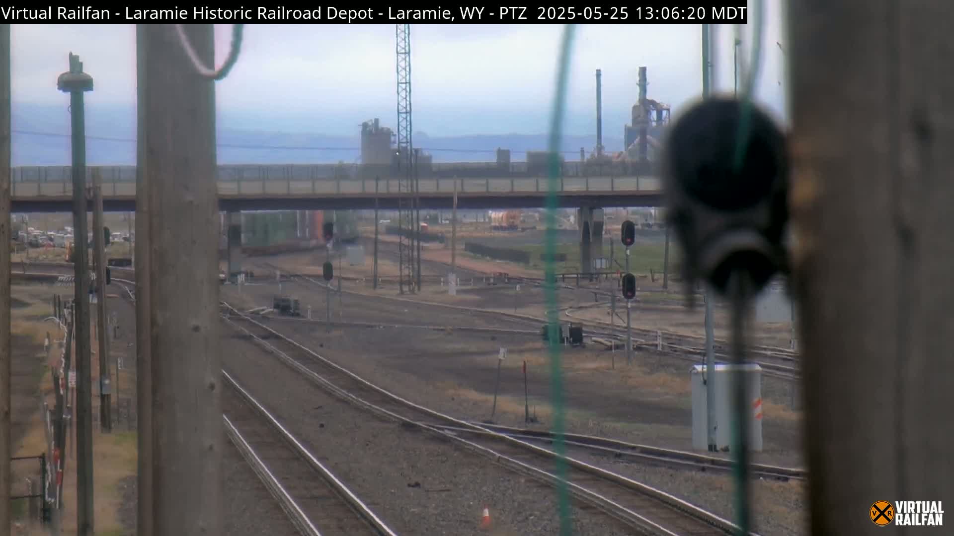 A view of multiple train tracks intersecting under an overpass on an overcast day, with an industrial plant visible in the background.
