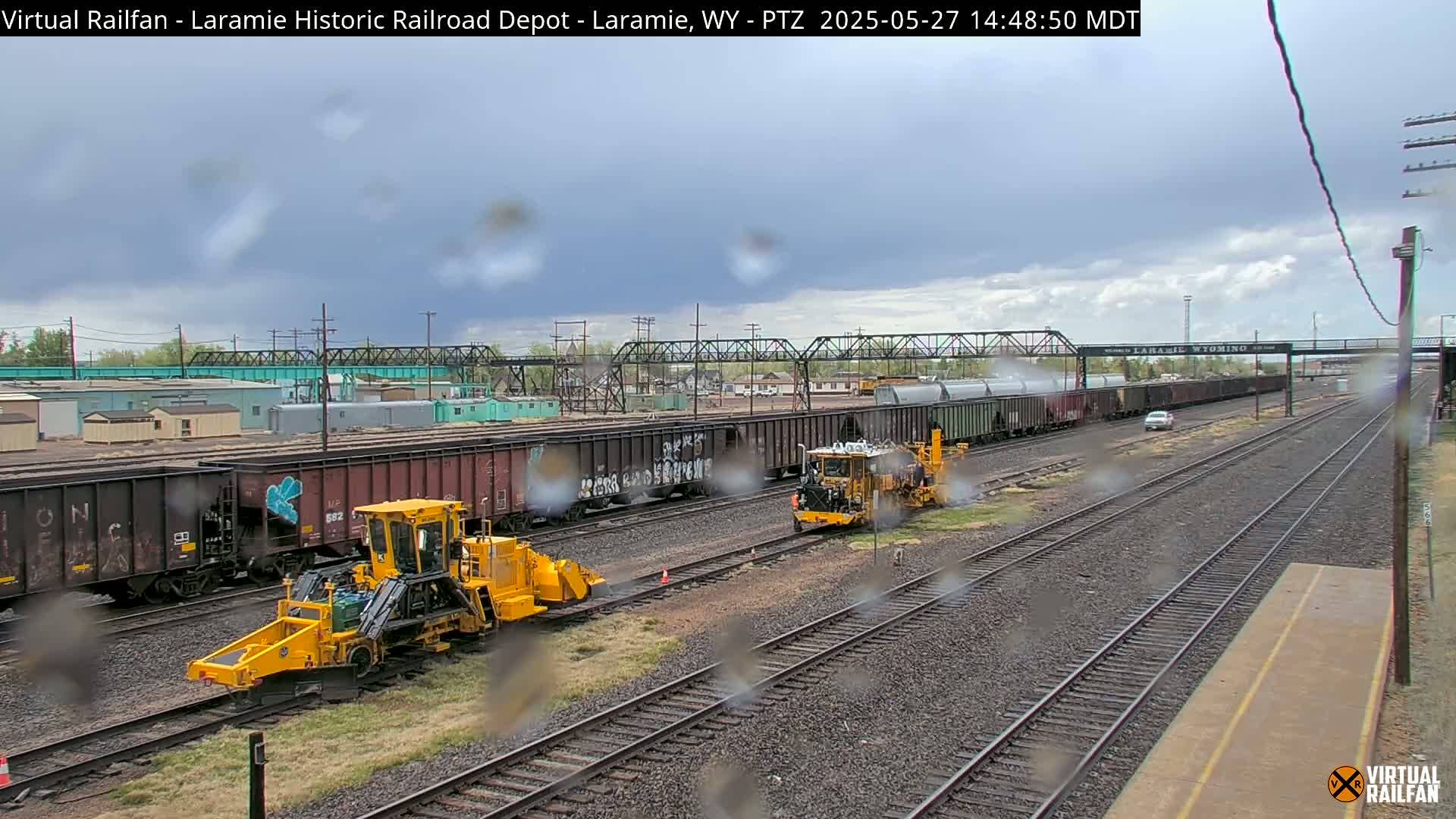 Two yellow rail maintenance vehicles are positioned on parallel train tracks under an overcast sky, with rain visible on the camera lens, alongside several freight cars.