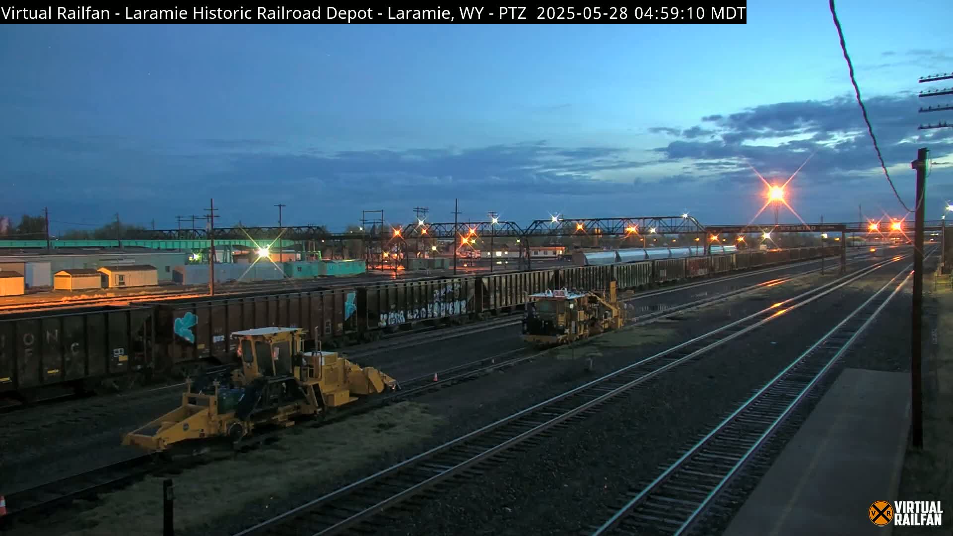 Under a twilight sky, two yellow rail maintenance machines are positioned along multiple train tracks beside a rail yard with several freight cars.