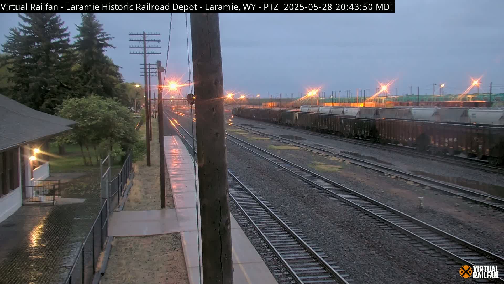 A long freight train sits on a set of tracks next to a depot building under a dim, possibly rainy, evening sky.