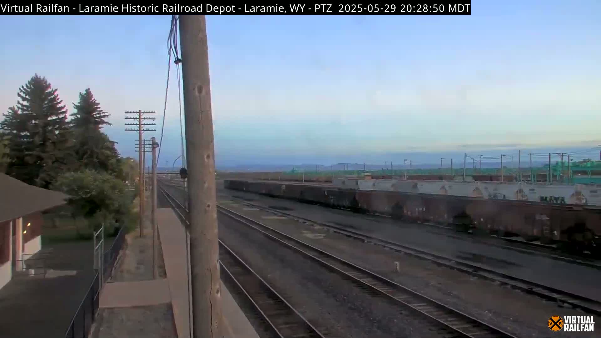 Under a clear, twilight sky, a long line of freight cars sits on a rail yard next to a set of parallel train tracks and a small building.