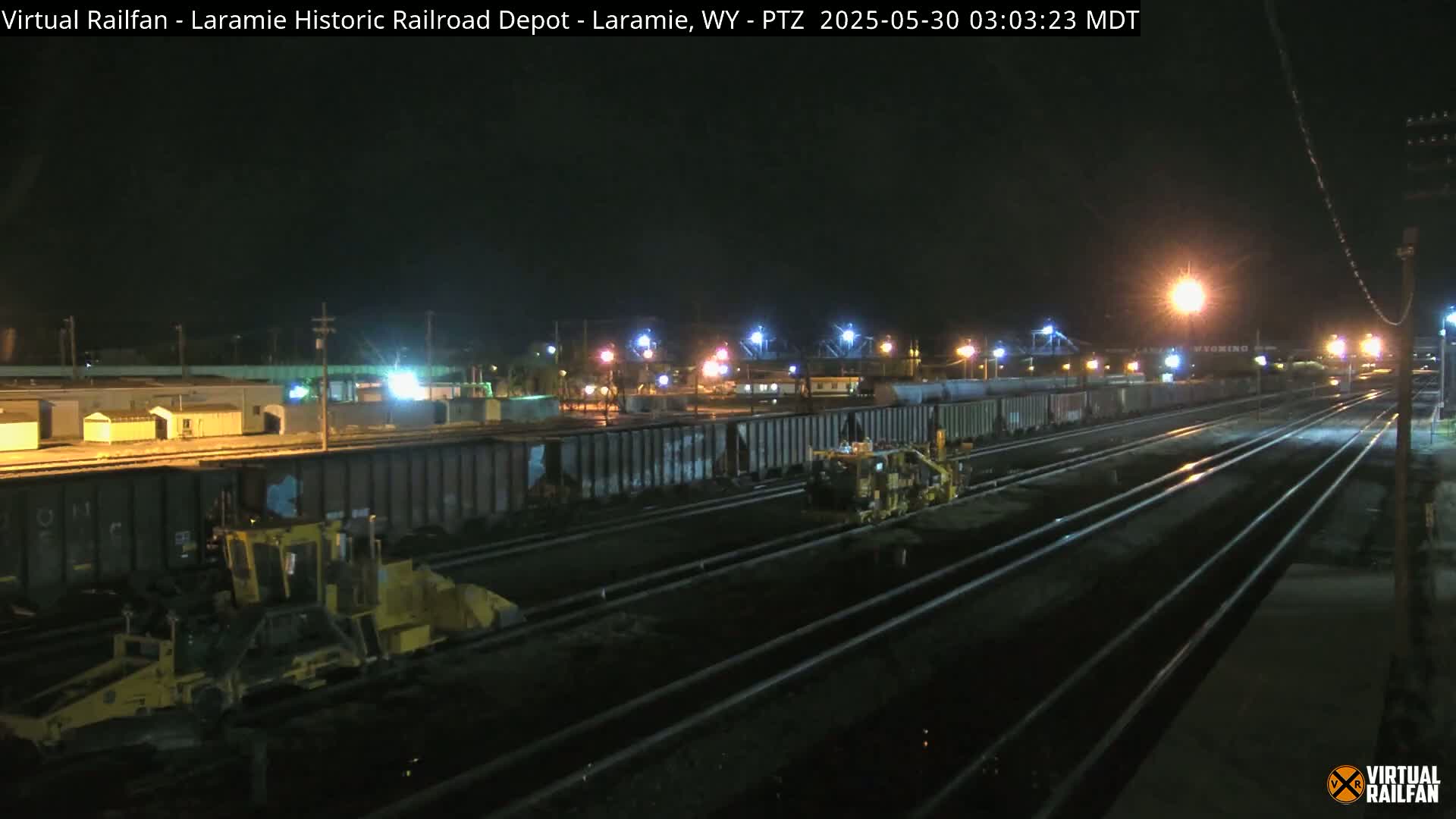 At night, two yellow pieces of heavy machinery are working on a train track surrounded by many dark rail cars under bright lights.