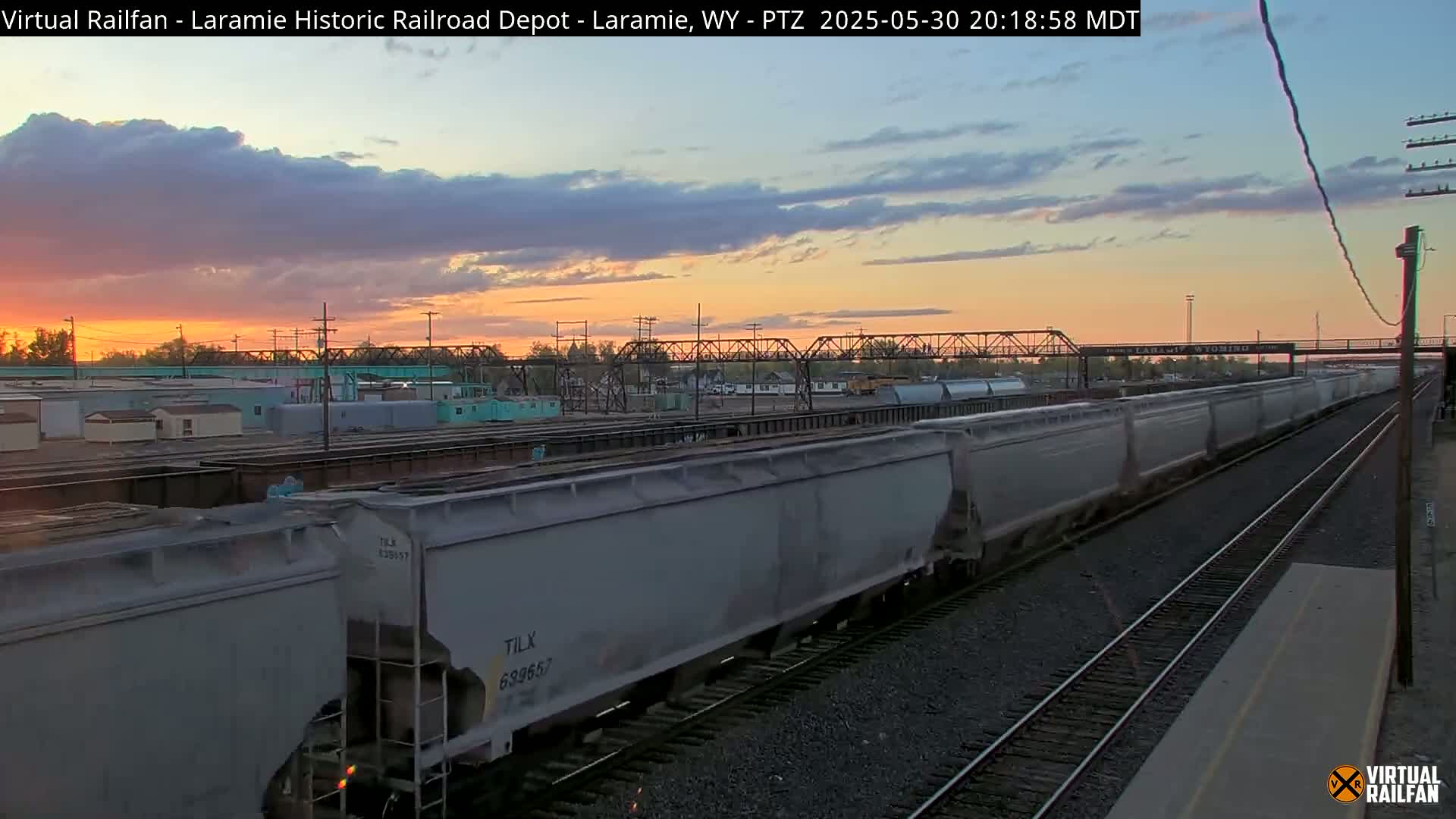 A long freight train of gray cars moves along railroad tracks during a sunset with a mostly cloudy sky.