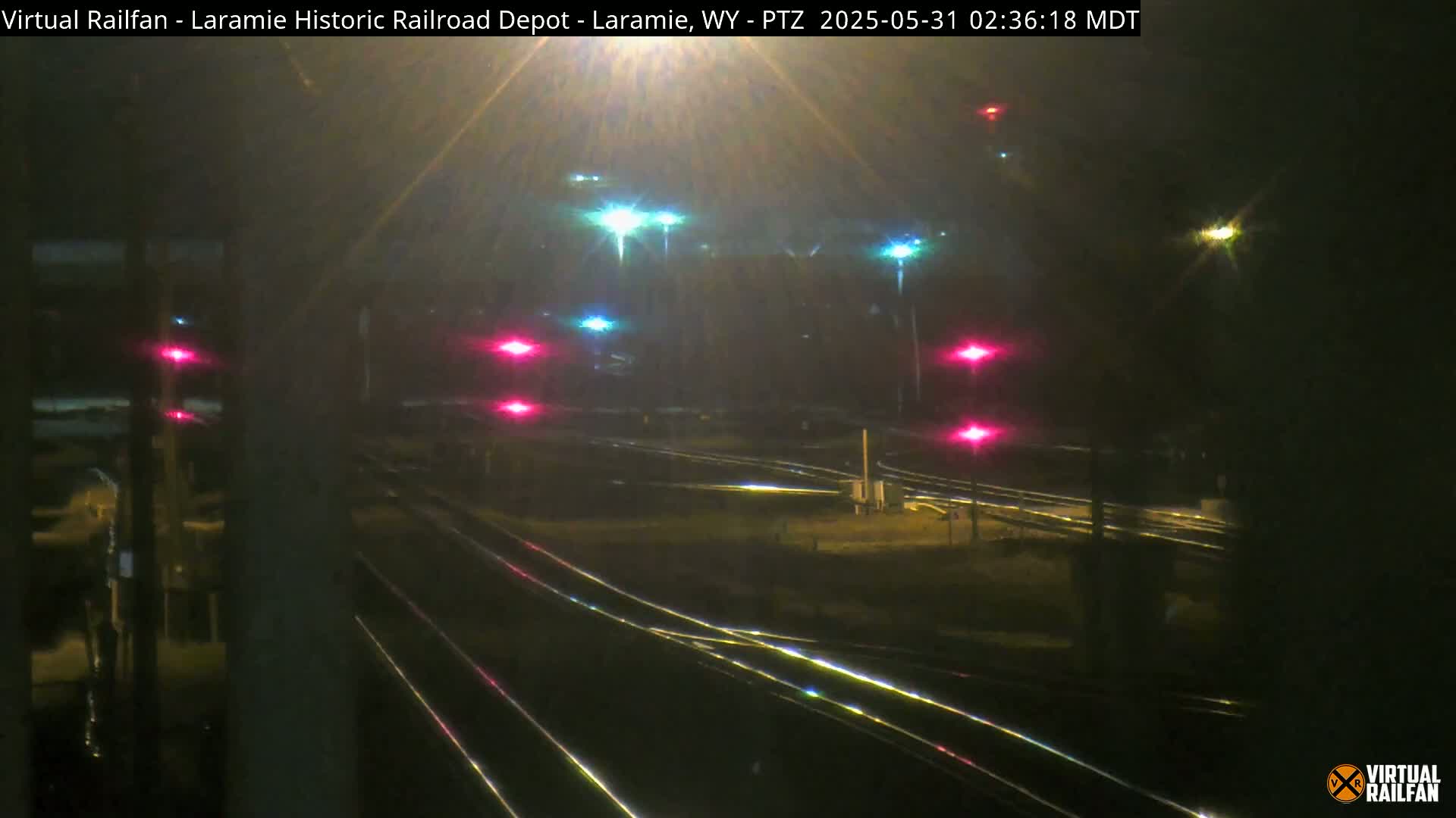 A nighttime view of a railroad yard with multiple sets of tracks, illuminated by various lights, some of which appear red.