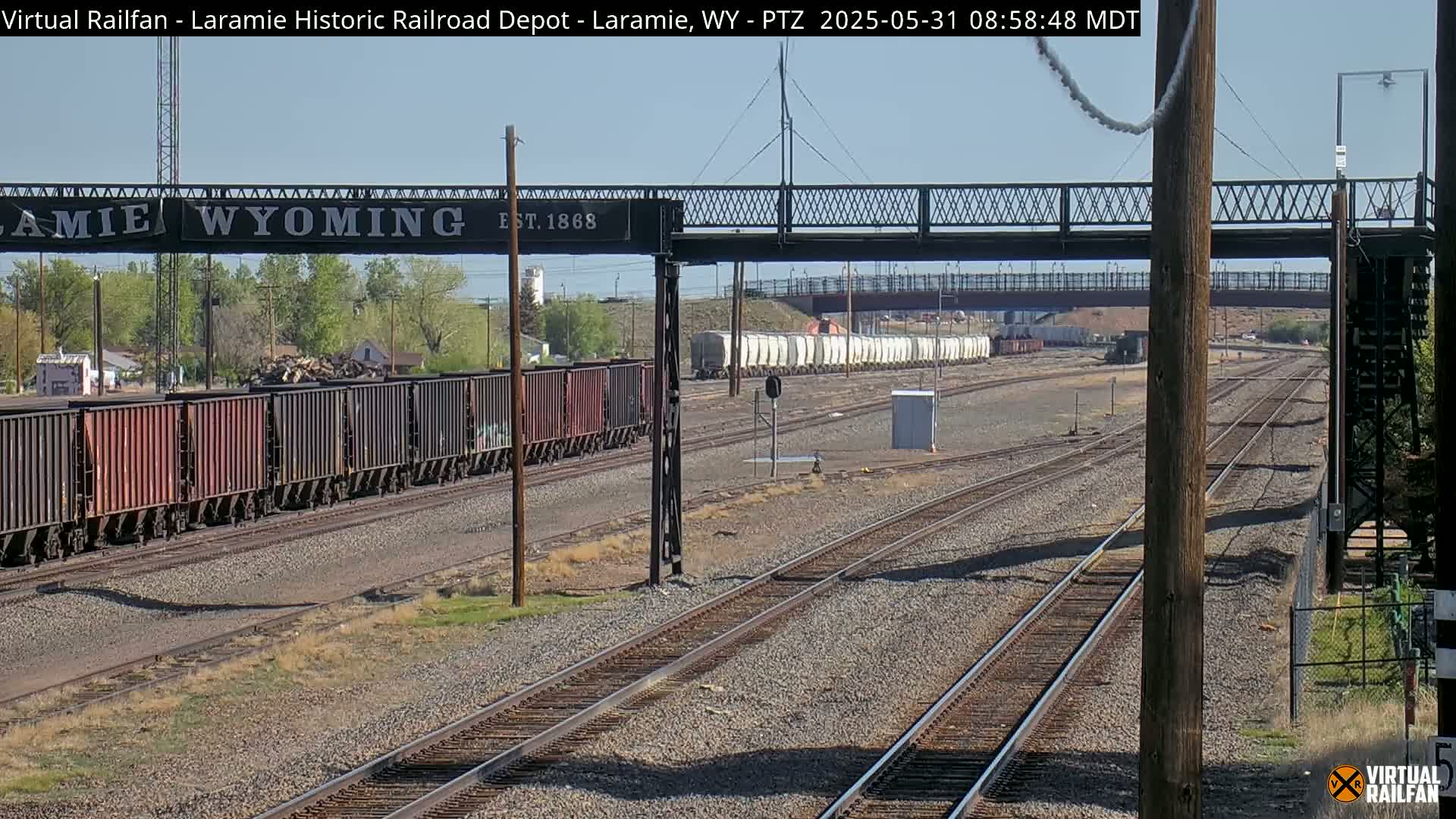 Under a clear sky, several sets of railroad tracks stretch into the distance, with freight cars and a pedestrian bridge visible.