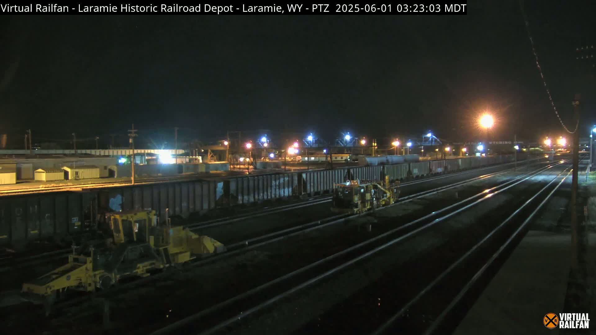 At night, under artificial lights, several pieces of heavy machinery are working alongside a long line of dark train cars sitting on railroad tracks.