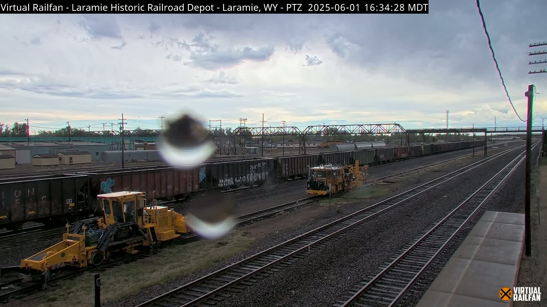Under a cloudy sky, a railway yard shows several freight cars and two pieces of yellow railway maintenance equipment near multiple sets of tracks.