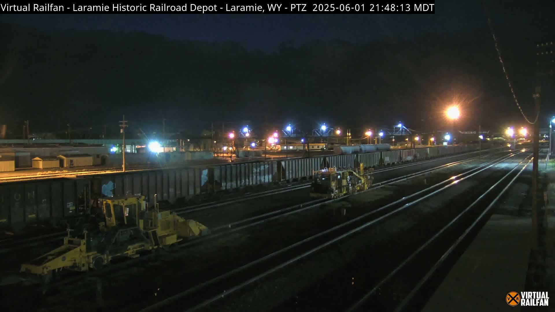 At night, under a dark sky, two pieces of yellow railway maintenance equipment are positioned near a train of dark railcars on a set of railroad tracks.