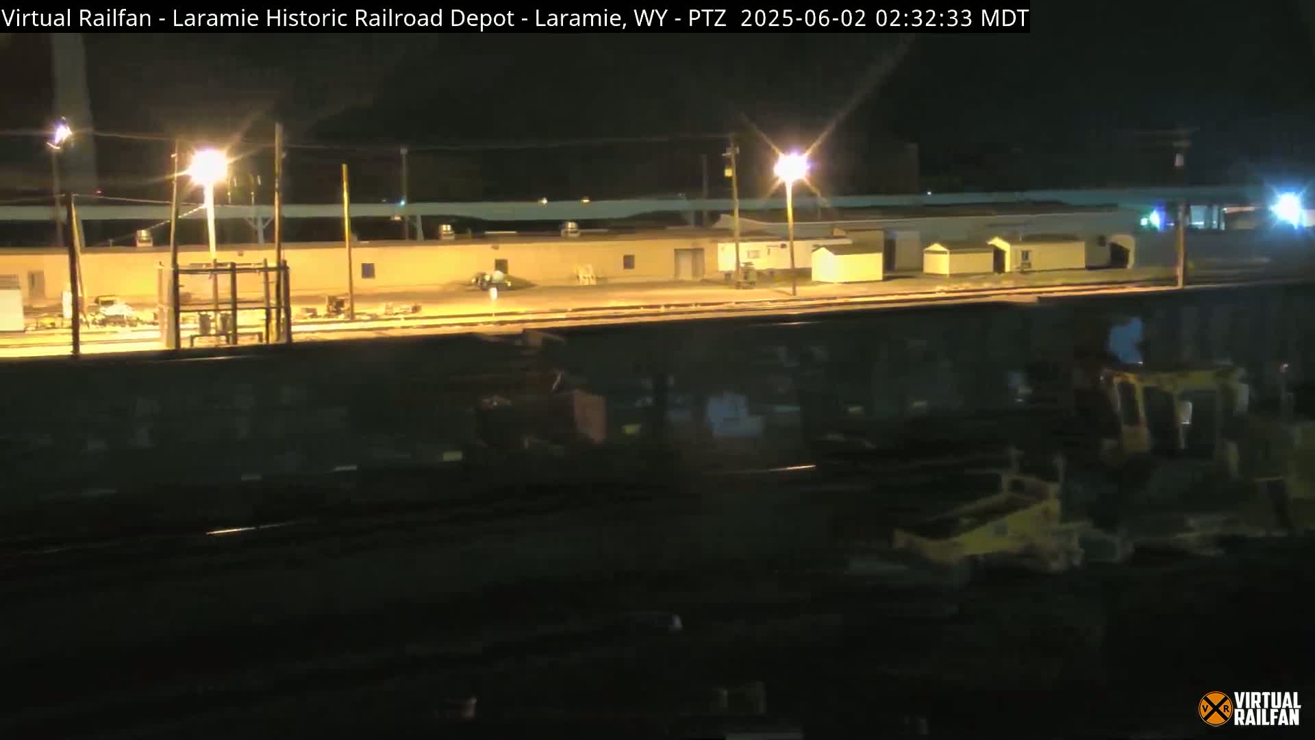 At night, a rail yard is illuminated by several lights, showing a large, dark railcar and a piece of yellow construction equipment.