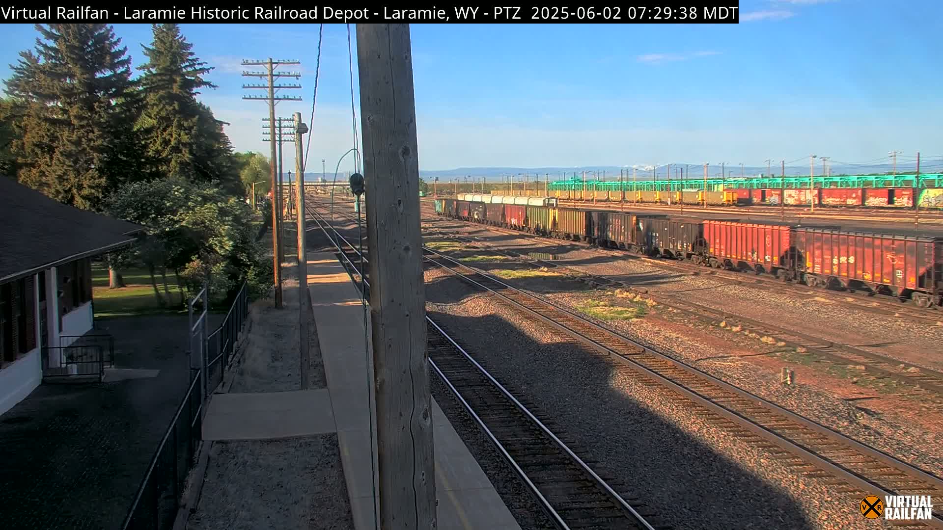 A sunny day reveals a railroad yard with multiple train cars, tracks, and a small building next to a grassy area with trees.