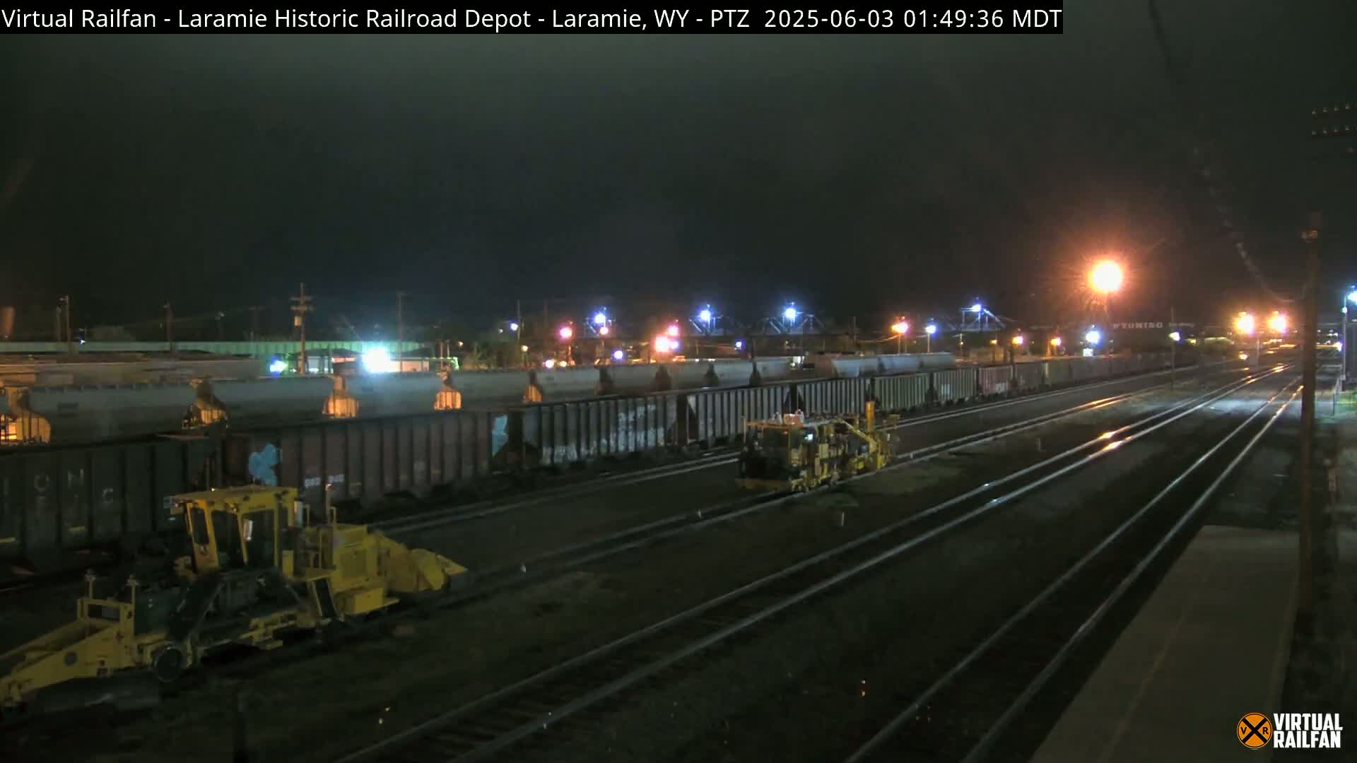 At night, a long line of rail cars sits on tracks next to two pieces of yellow maintenance equipment.