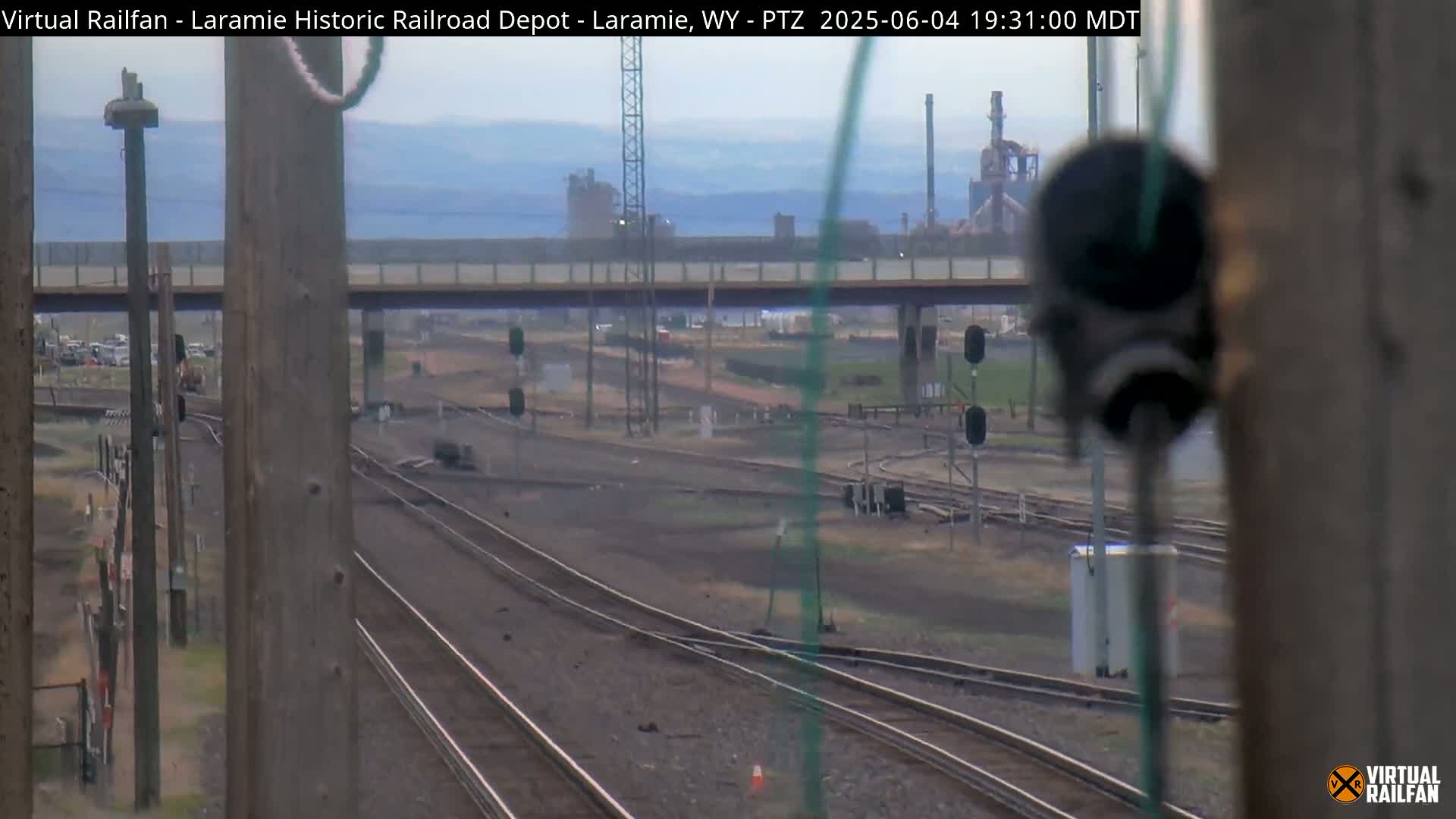 Multiple sets of train tracks extend into the hazy distance, passing under a bridge and toward an industrial area on an overcast day.