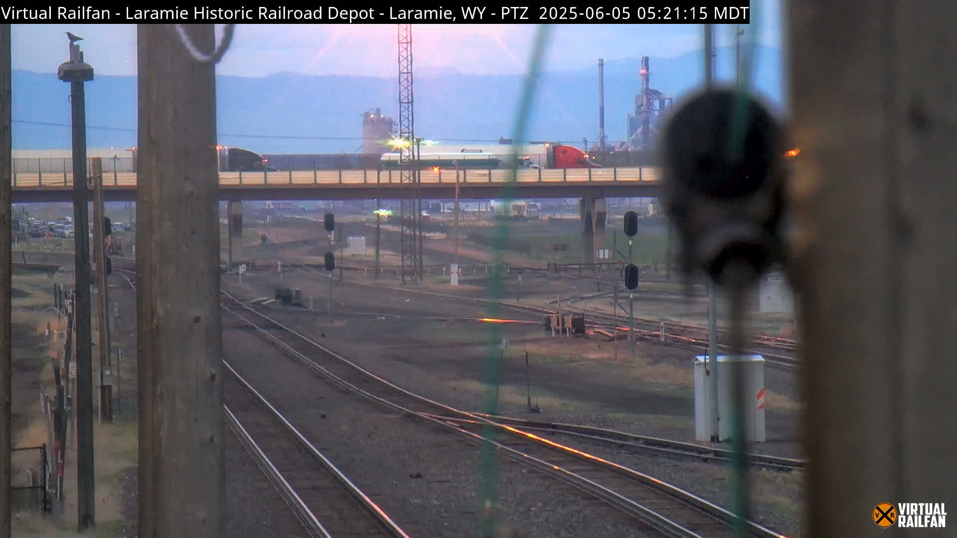 A view of railroad tracks and a highway overpass at sunrise, with industrial buildings in the distance.