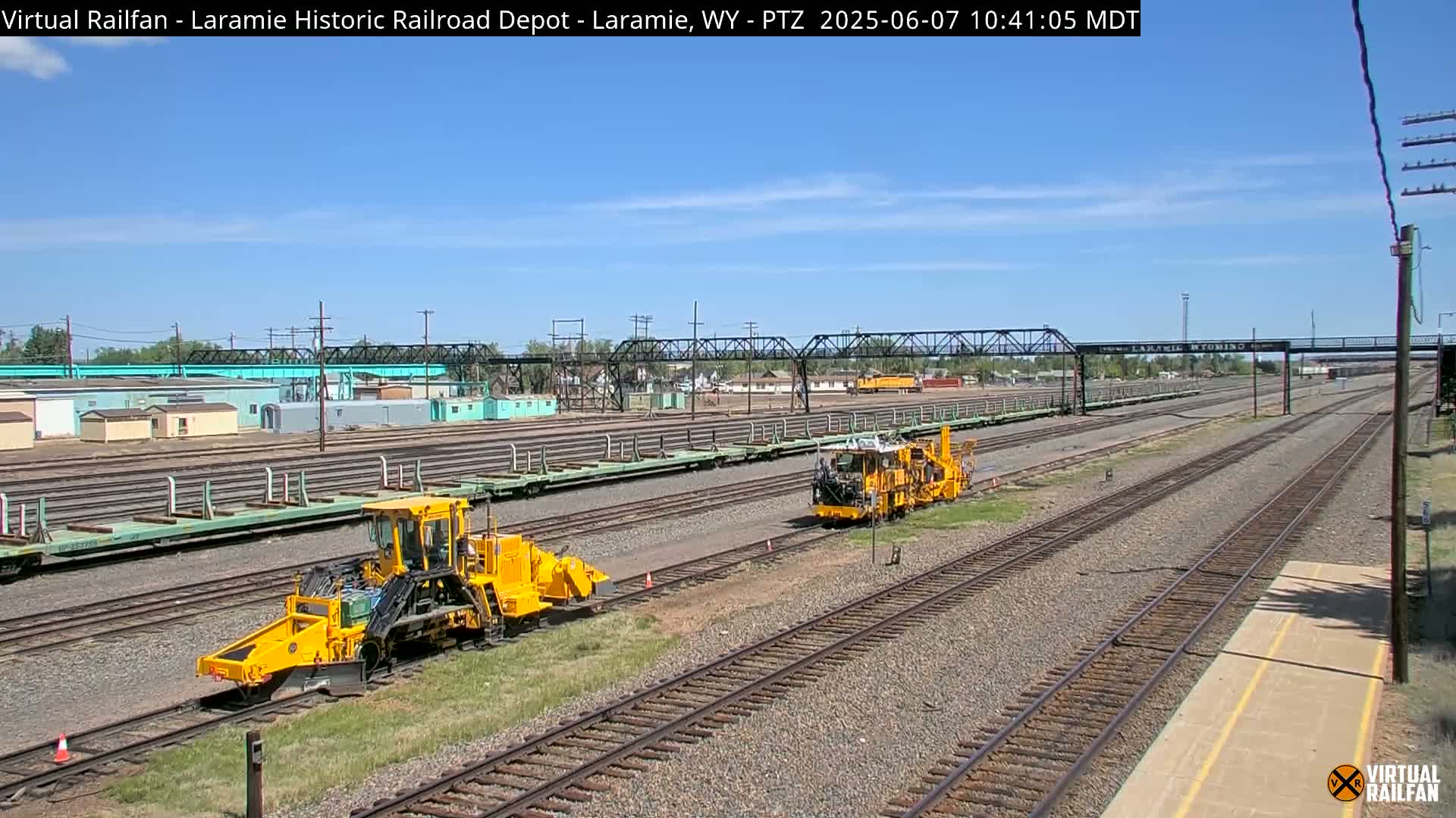 Under a clear blue sky, two yellow rail maintenance vehicles are positioned on a sprawling railroad yard with numerous tracks, alongside light-green rail cars and buildings.
