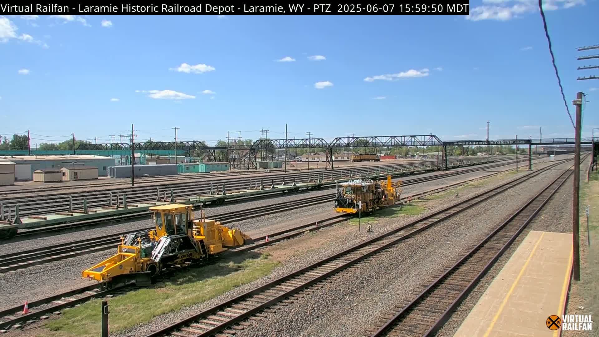 Under a mostly sunny sky, two yellow rail maintenance vehicles are positioned on a large rail yard with numerous parallel train tracks and several train cars.