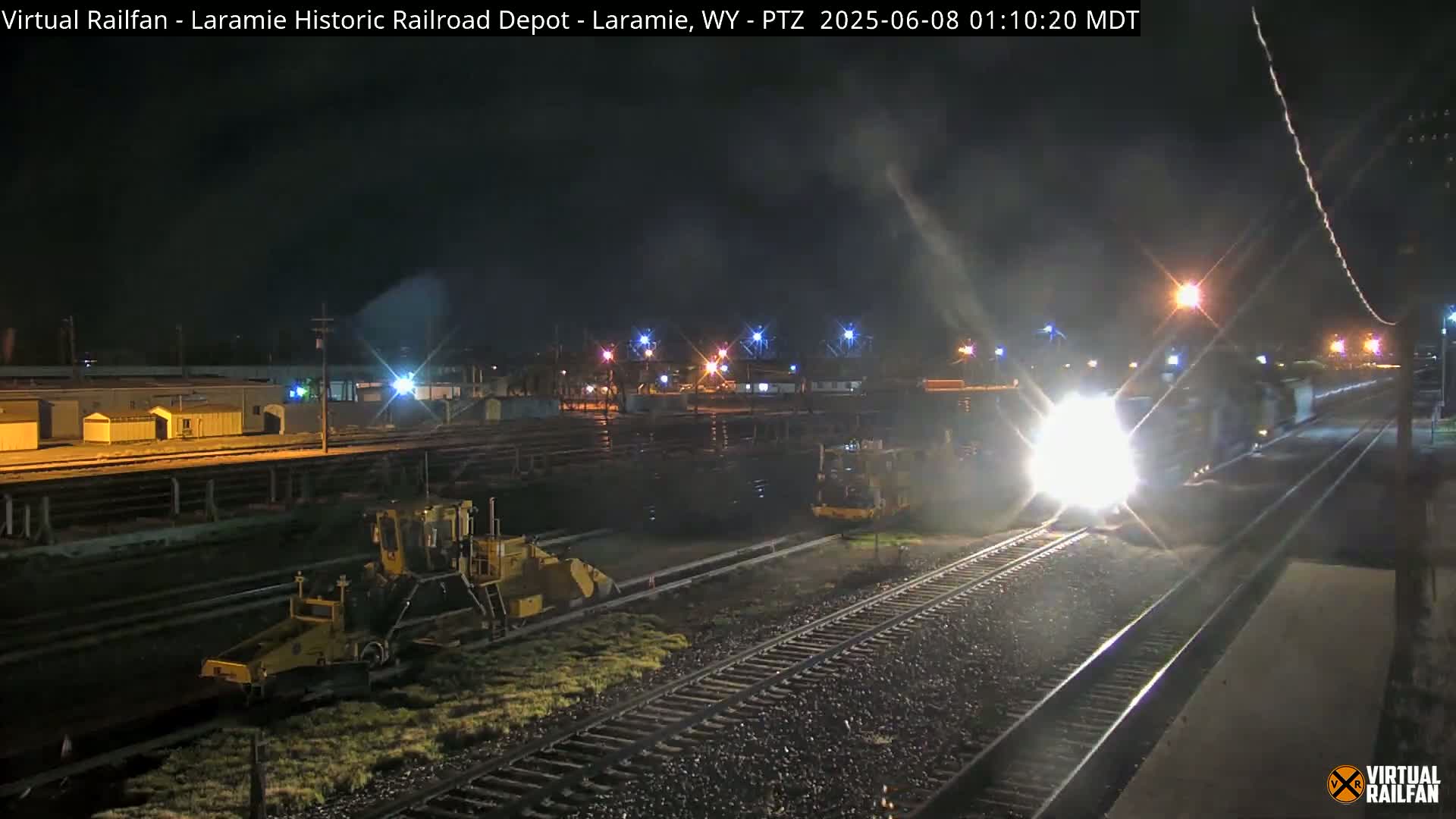At night, a train approaches a railway depot where two pieces of track maintenance equipment are parked alongside the tracks.