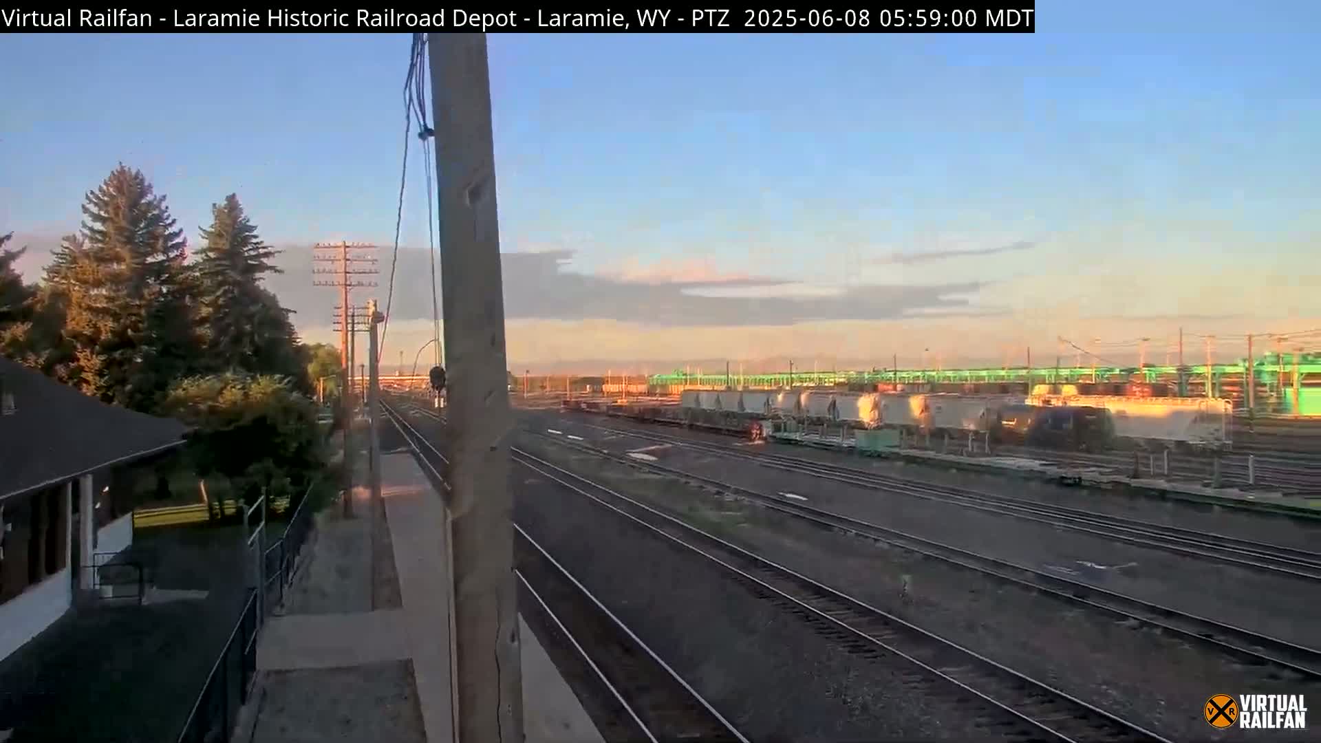 A long shot of a train yard at sunrise, showing several rail lines and a variety of freight cars, with trees and a small building visible in the foreground.