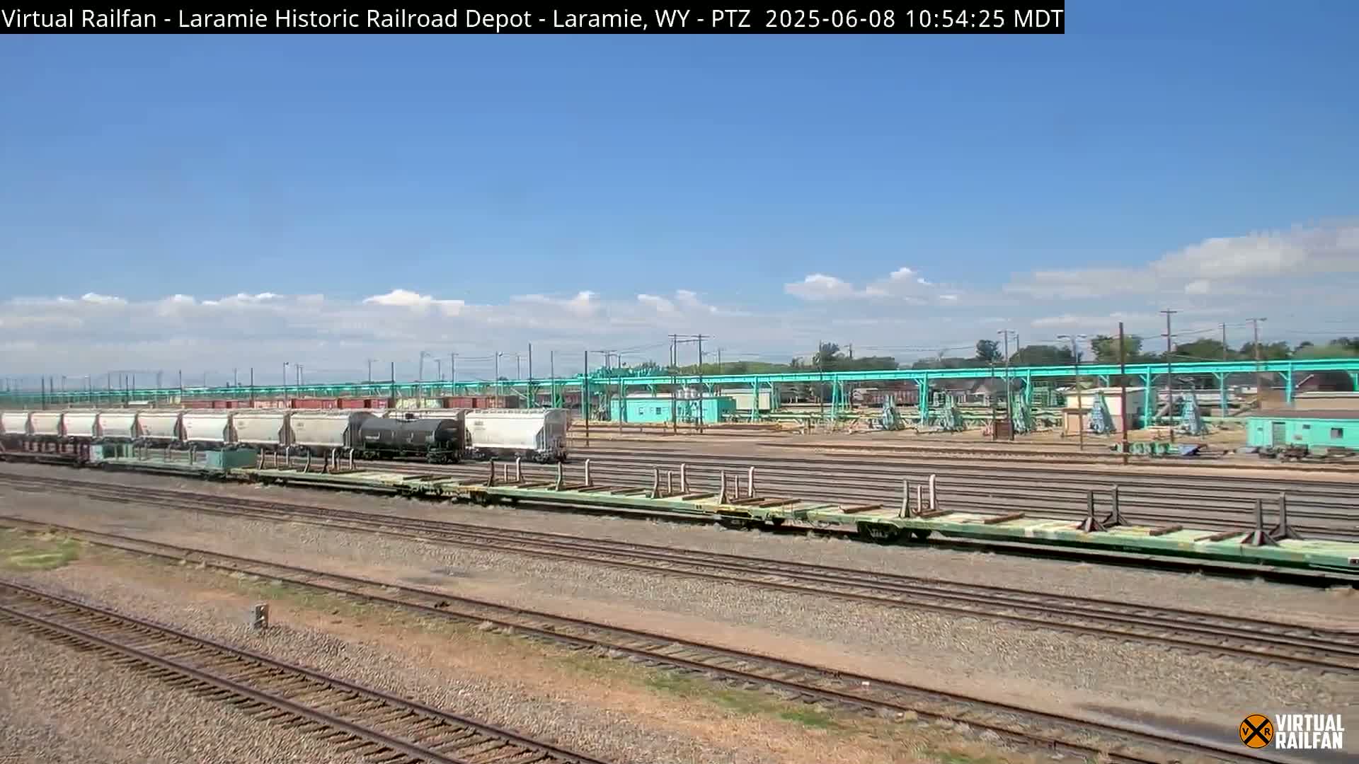 A long freight train sits on a rail yard under a mostly sunny sky with a few scattered clouds.
