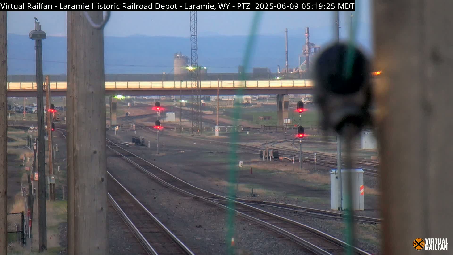 Multiple sets of railroad tracks converge under a bridge on a mostly clear day, with industrial structures visible in the distance.