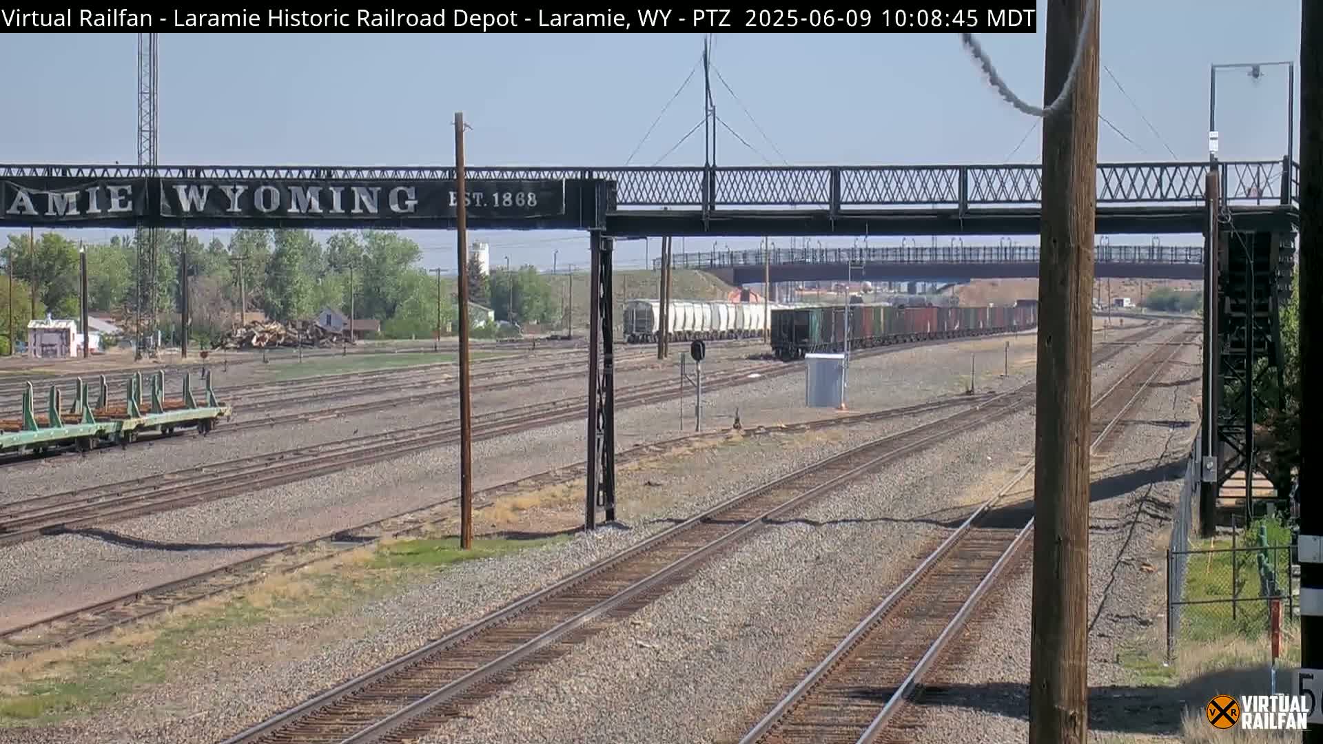 A train of light-colored boxcars passes under a metal footbridge on a sunny day, with multiple sets of train tracks extending into the distance.