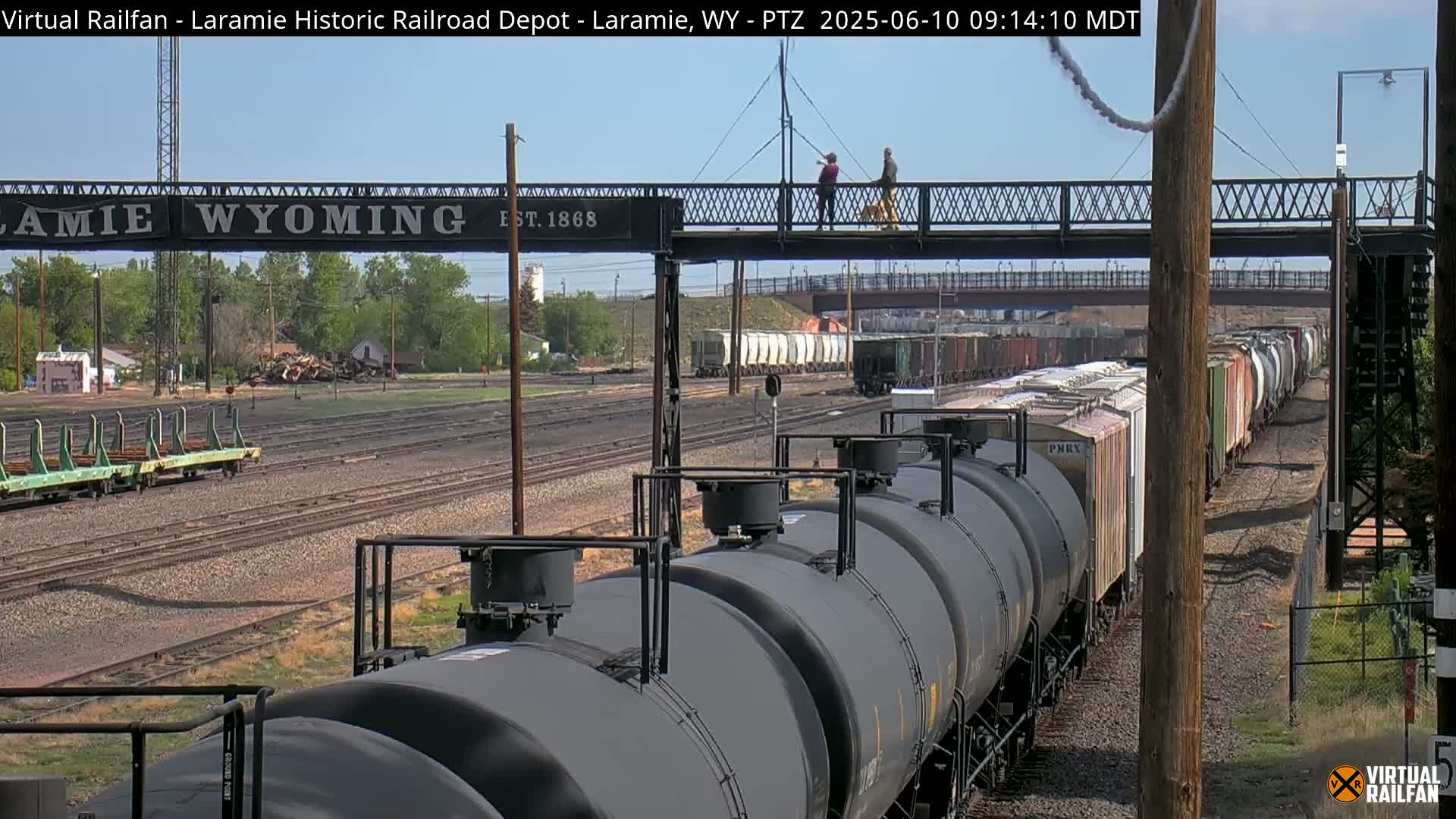 A long train of dark-colored tanker cars is seen on a rail line under a mostly sunny sky, with a pedestrian bridge and other railroad cars visible in the background.