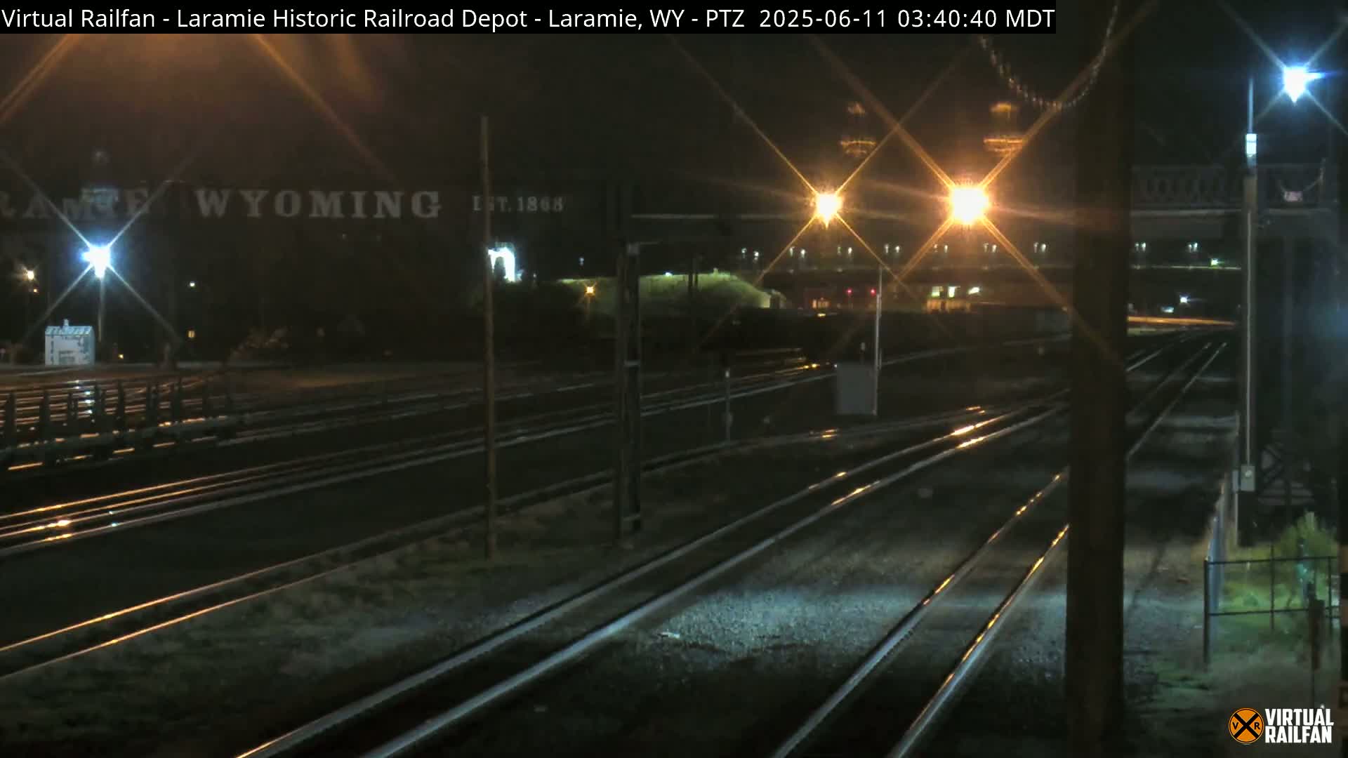 At night, several train tracks stretch into the distance under bright lights, past a building with "Laramie Wyoming Est. 1868" on its side.