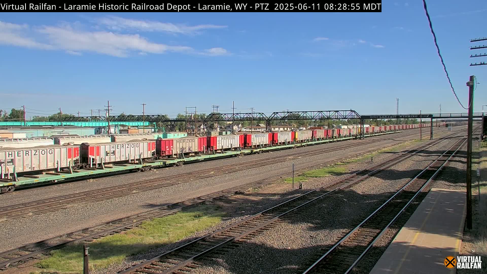 Under a mostly clear blue sky, a long freight train of covered hopper cars sits on a set of railroad tracks.