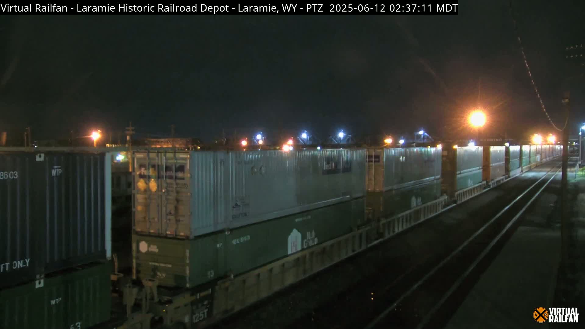 A long freight train carrying numerous shipping containers is visible at night under artificial lighting.