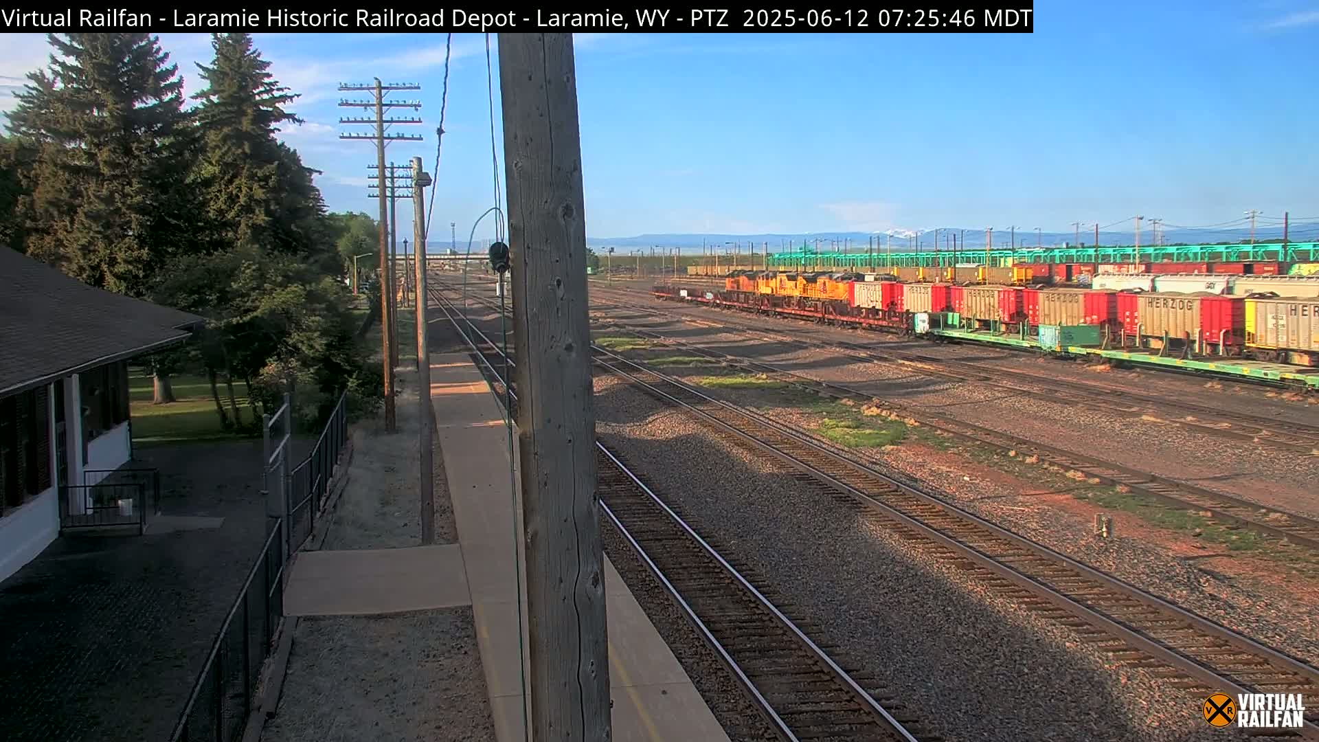 A sunny day reveals a train depot with a small building, green trees, and several rail cars parked on multiple tracks.