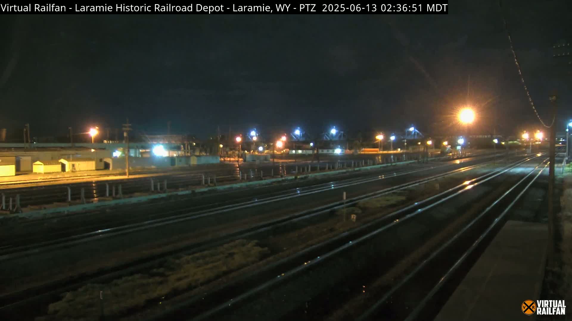 A nighttime view of multiple train tracks extending into the distance, wet from recent rain, with buildings and lights visible in the background.