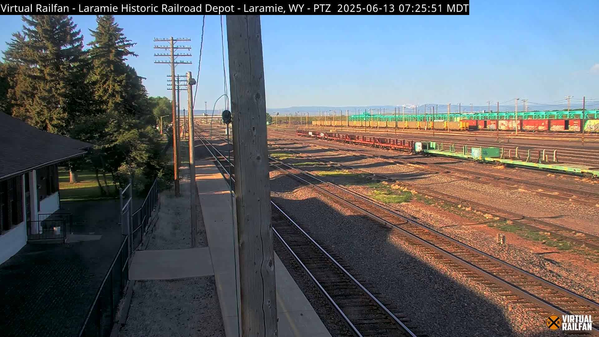 A sunny day reveals a railway yard with numerous train cars and tracks, alongside a small building and trees.