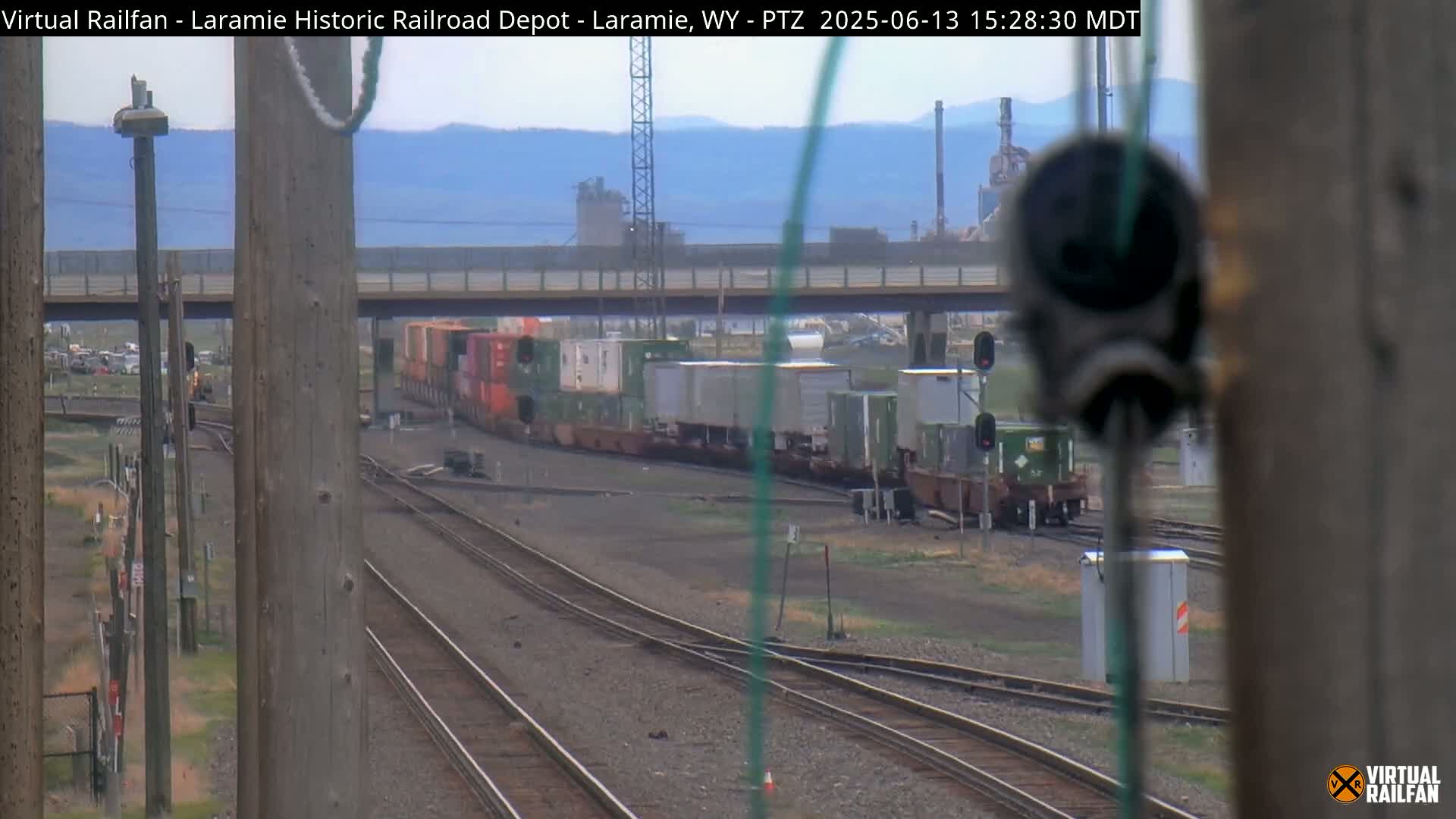 A long freight train is passing under a bridge on a partly cloudy day, with industrial buildings visible in the distance.