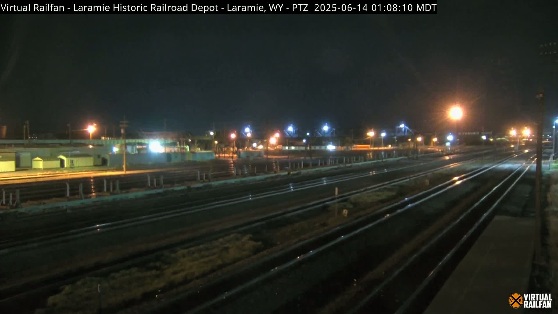 A nighttime view of a railroad yard with multiple tracks, illuminated by artificial lights, shows some wetness on the tracks.