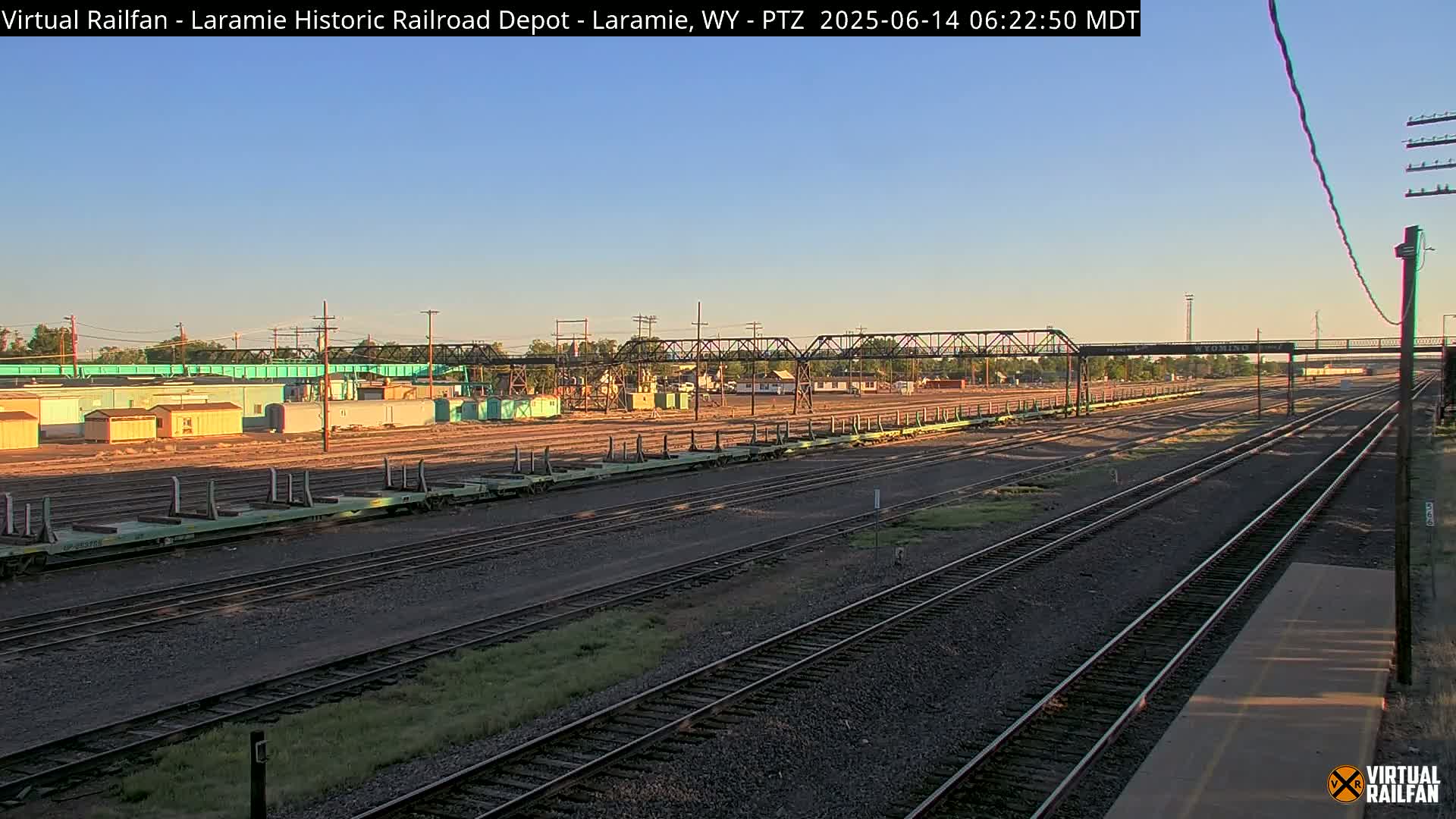 Under a clear morning sky, several parallel sets of railroad tracks extend into the distance, past a collection of railway cars and industrial buildings.