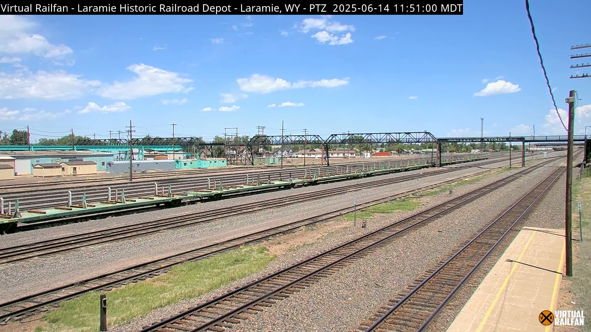 Several long, empty flatbed train cars sit on a set of railroad tracks under a mostly sunny sky.