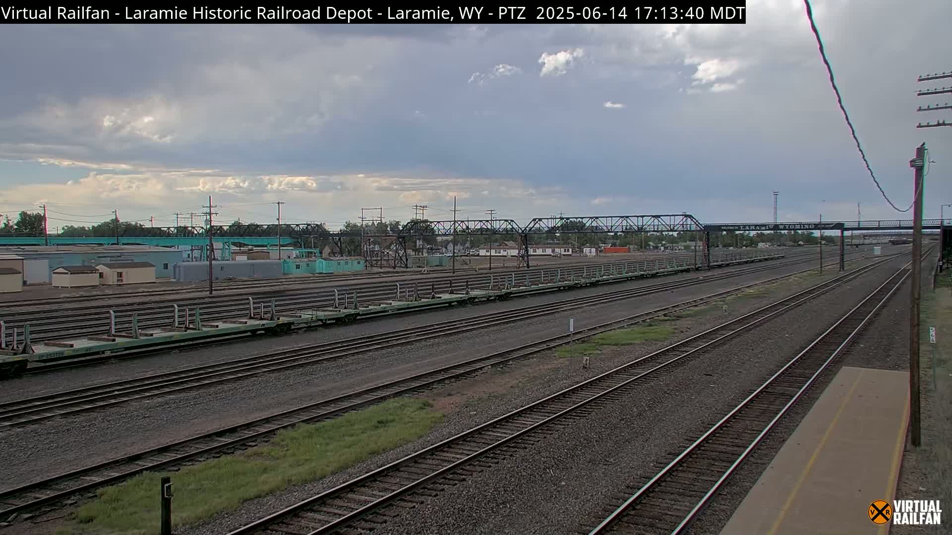 Several long, empty flatbed train cars sit on a set of railroad tracks under a mostly sunny sky.