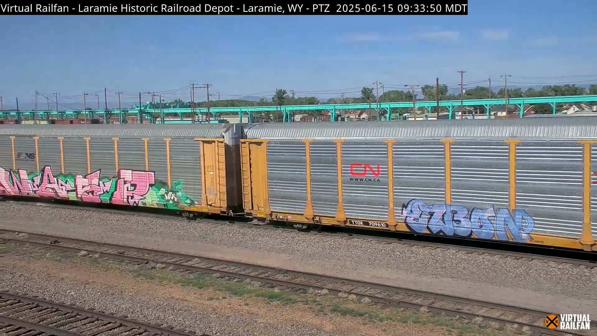 Two graffiti-covered freight train cars sit on a railway track under a clear, sunny sky.