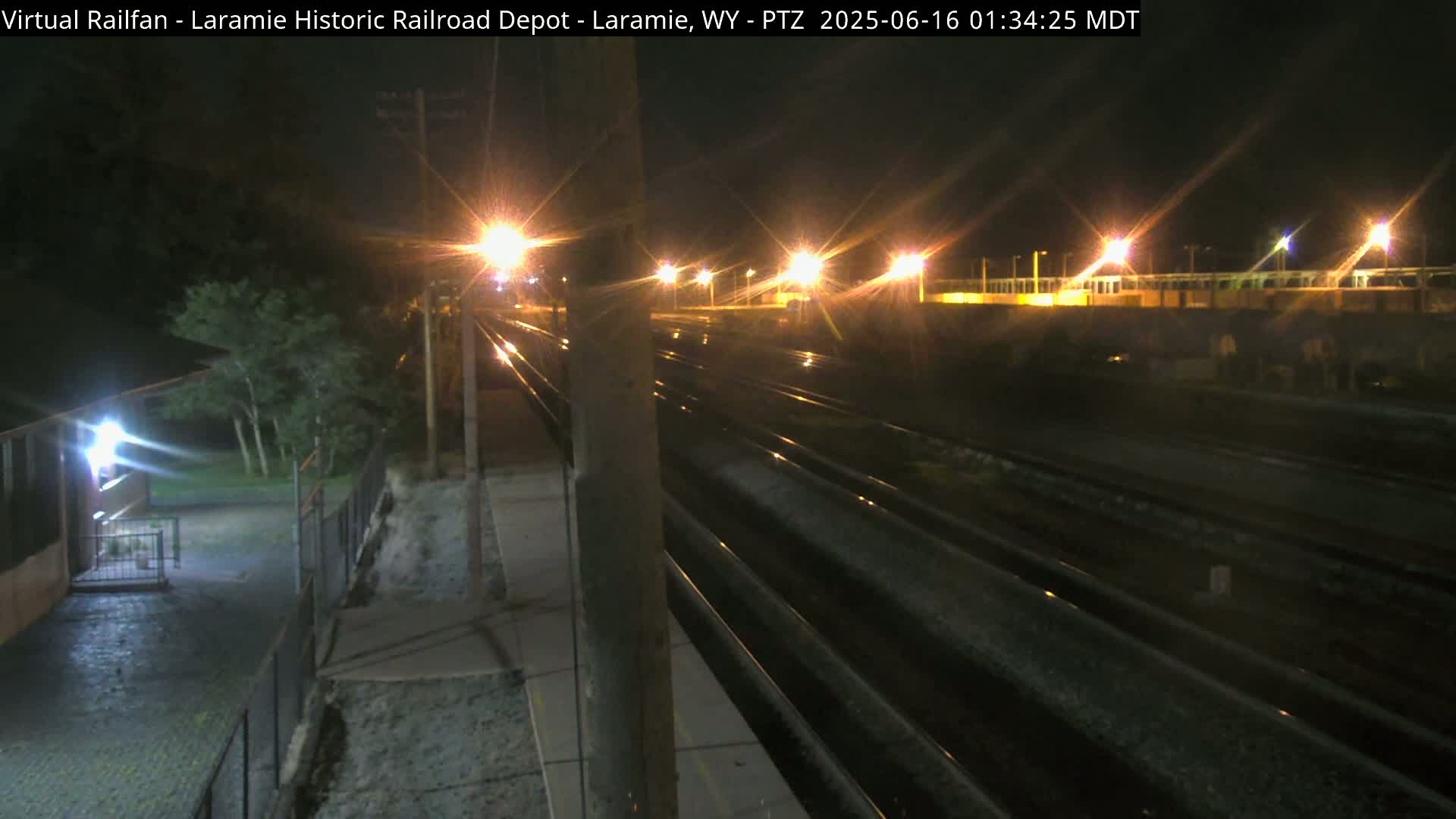 A nighttime view of wet railroad tracks extending into the distance, illuminated by streetlights, with a building and trees visible in the foreground.