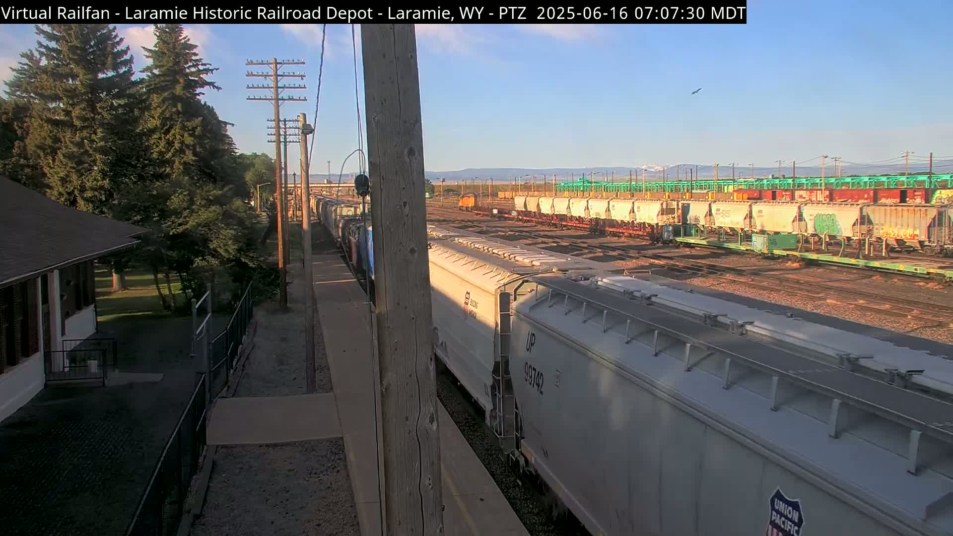 A long train of light-grey hopper cars passes by a smaller building and several more rail cars under a clear, sunny sky.