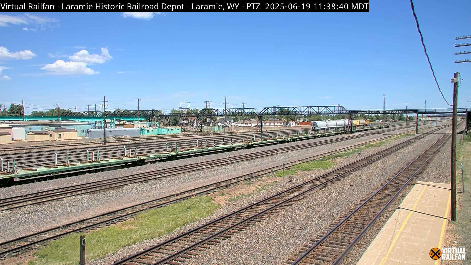 Under a partly cloudy blue sky, a long view of railroad tracks shows several rail cars and a metal bridge in the distance.