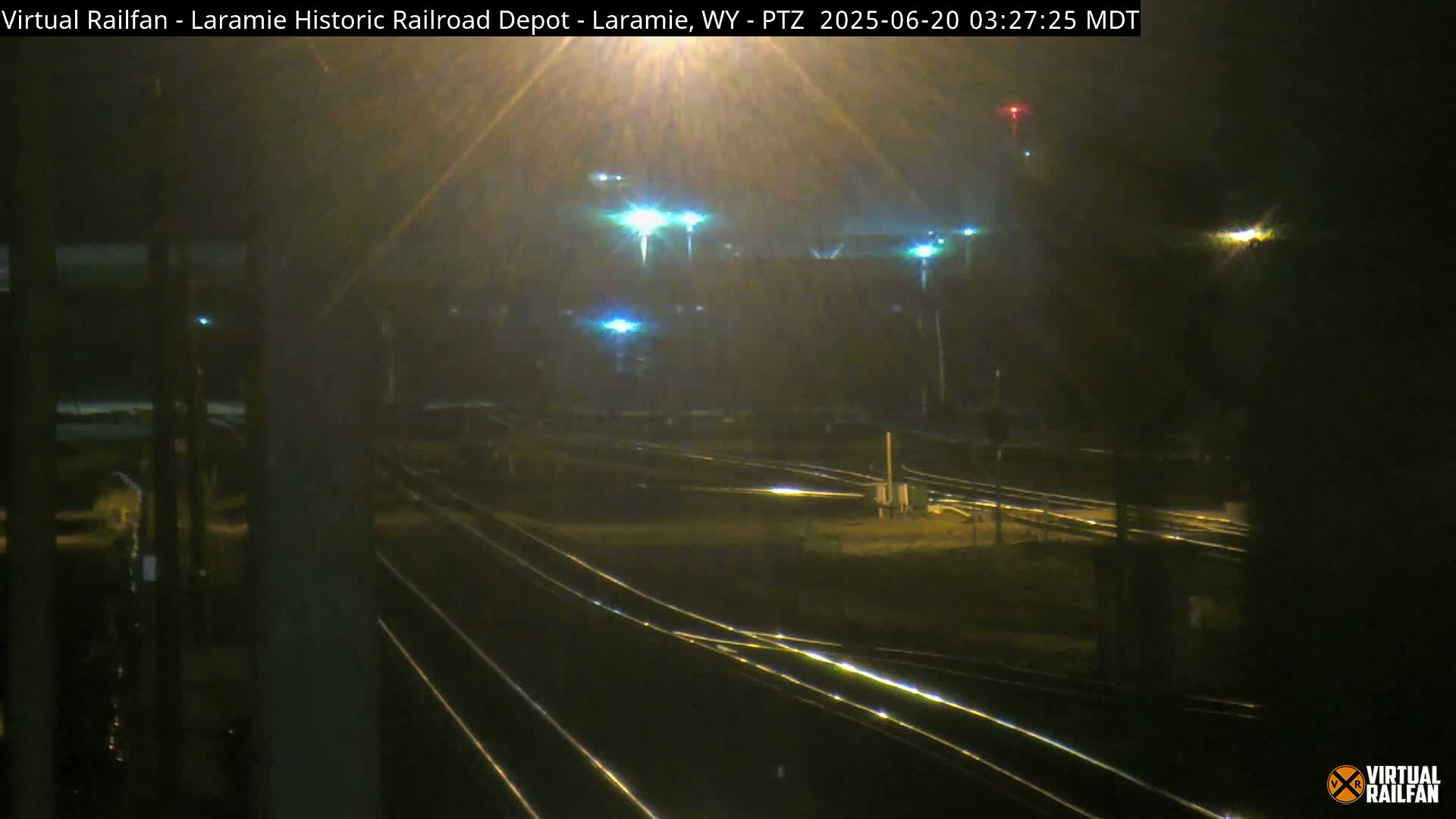 A nighttime view of railroad tracks and switching yards illuminated by several lights under hazy conditions.