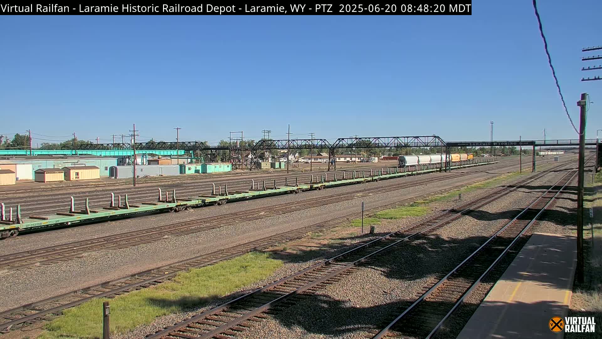 A long train of light green flatcars sits on a rail yard under a clear blue sky.