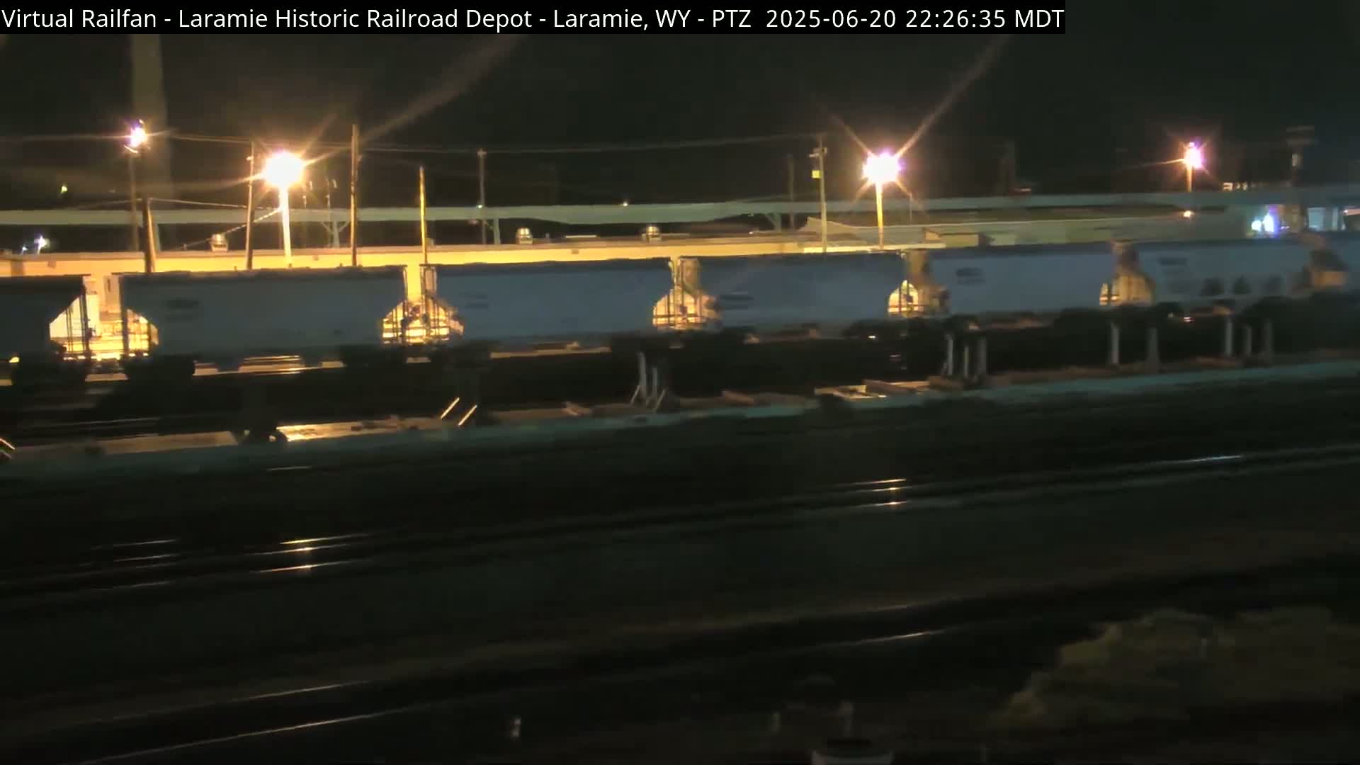 Several light-grey train cars sit on railroad tracks at night under artificial light.