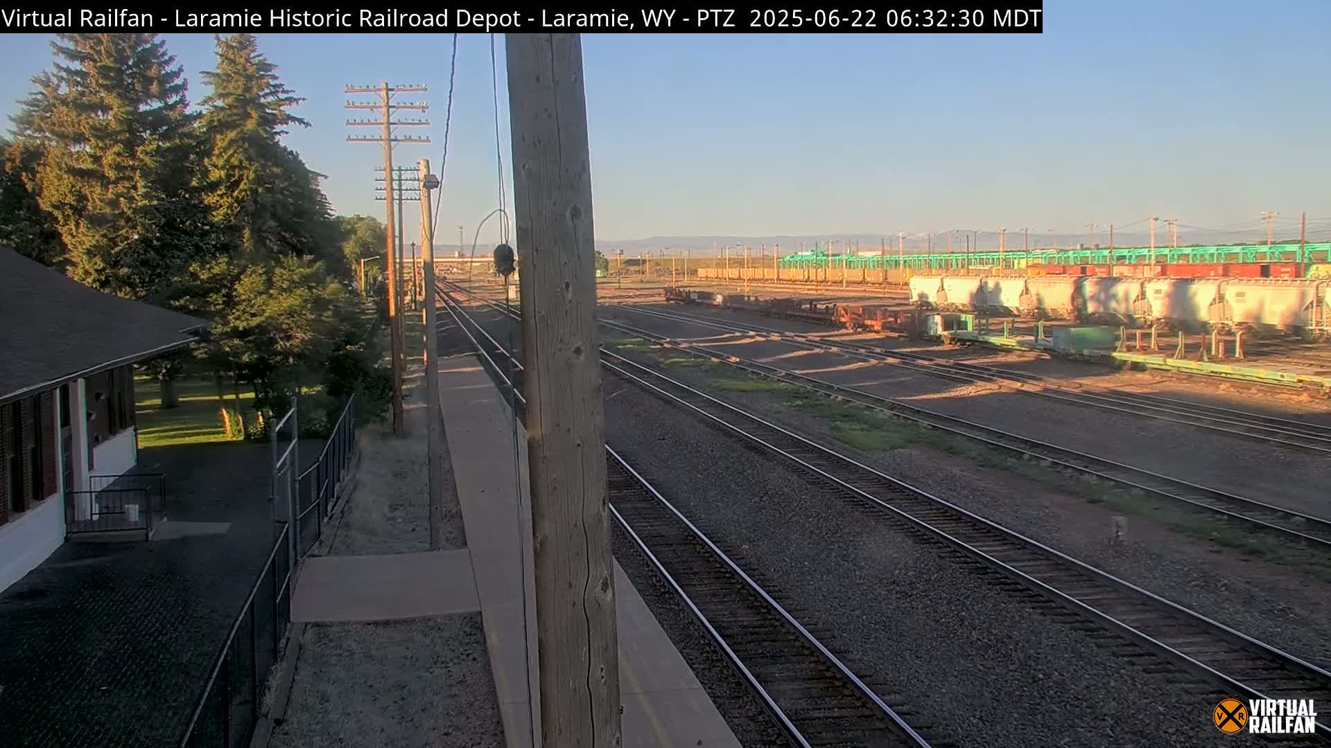 A railroad depot and several train cars sit on multiple parallel train tracks under a clear morning sky.