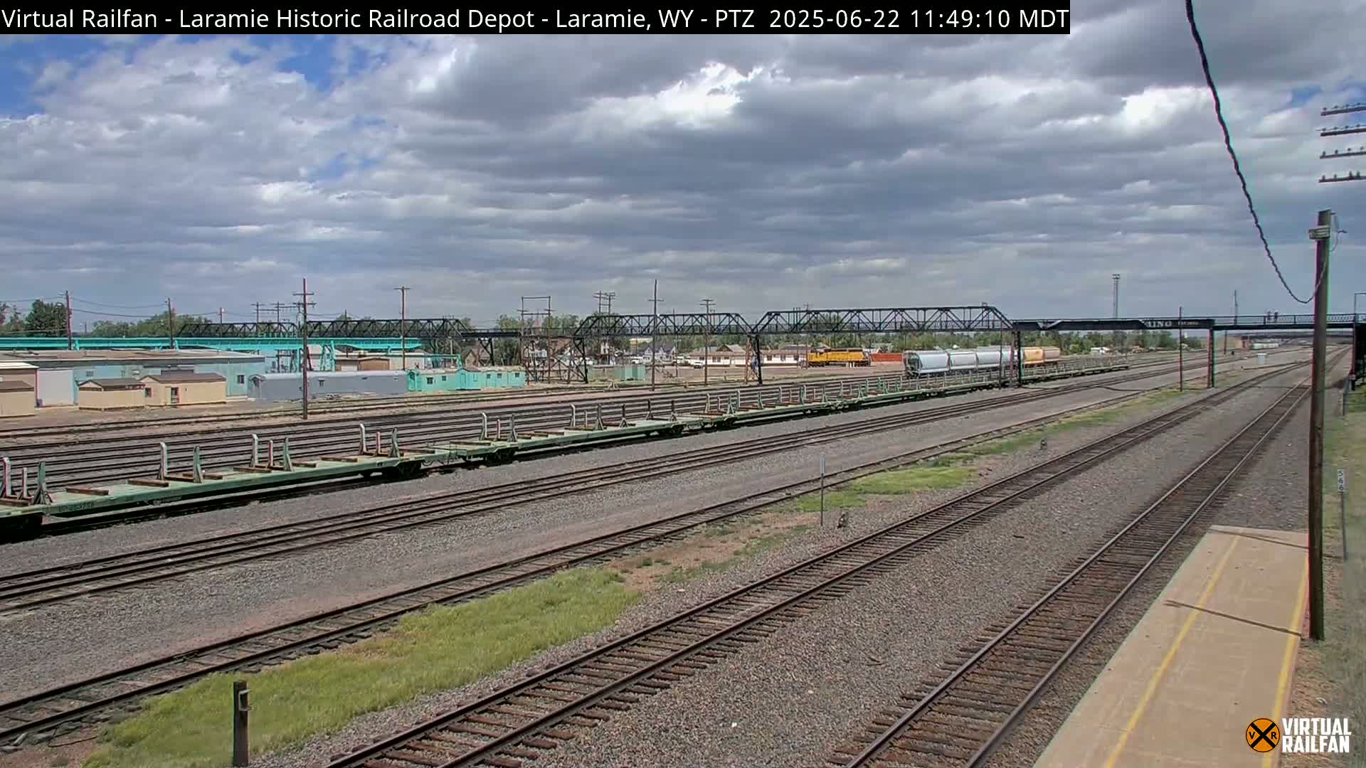 A cloudy day reveals a train yard with several rail cars and tracks stretching into the distance under a steel bridge.