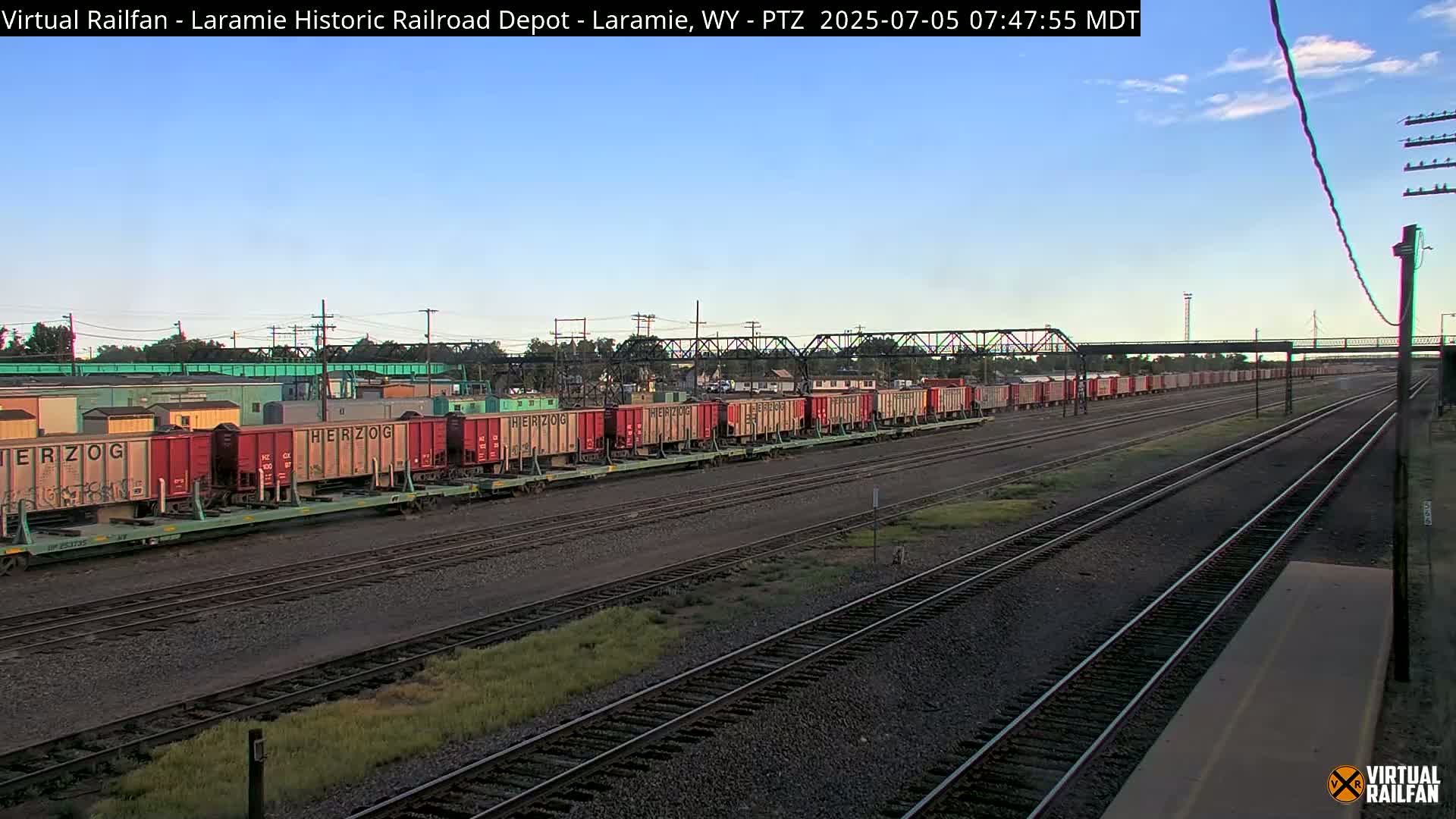 A long freight train of red and gray boxcars sits on a railway track under a clear blue sky.