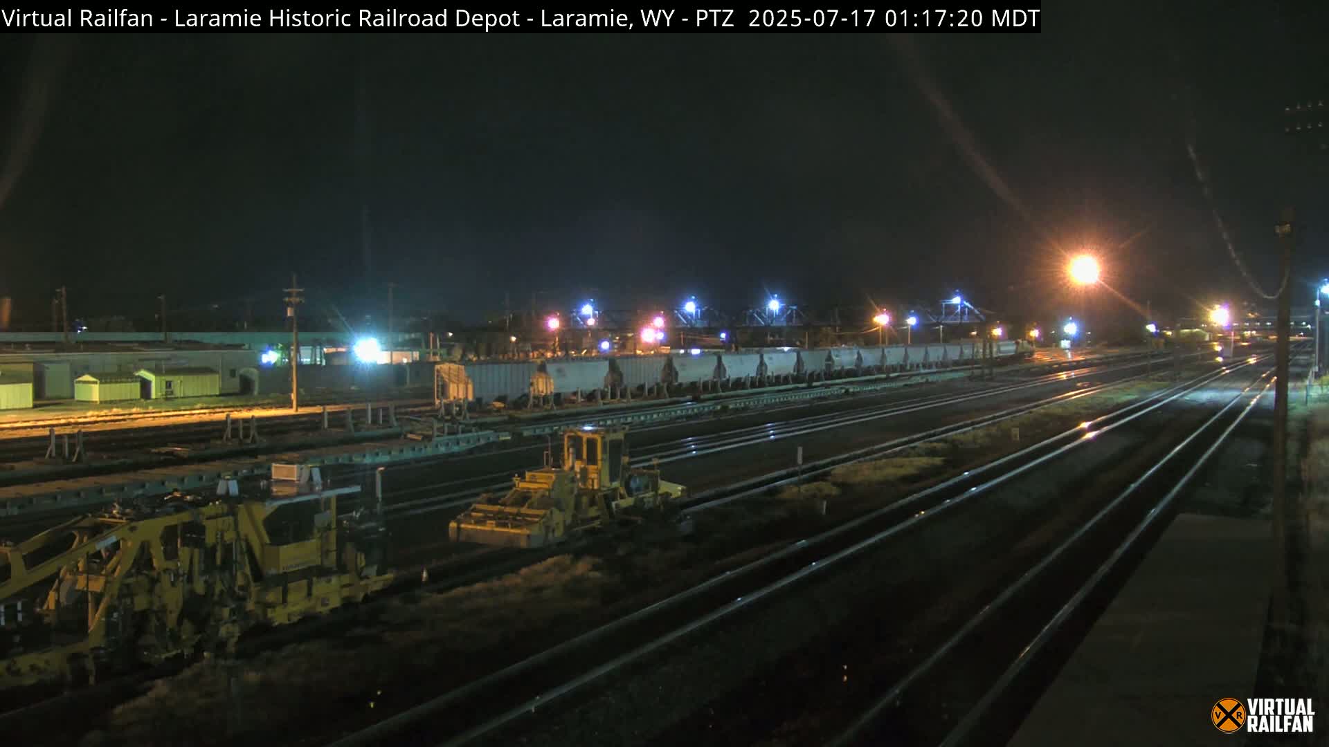 At night, under artificial lights, railroad maintenance vehicles are working near a long line of rail cars on multiple tracks.