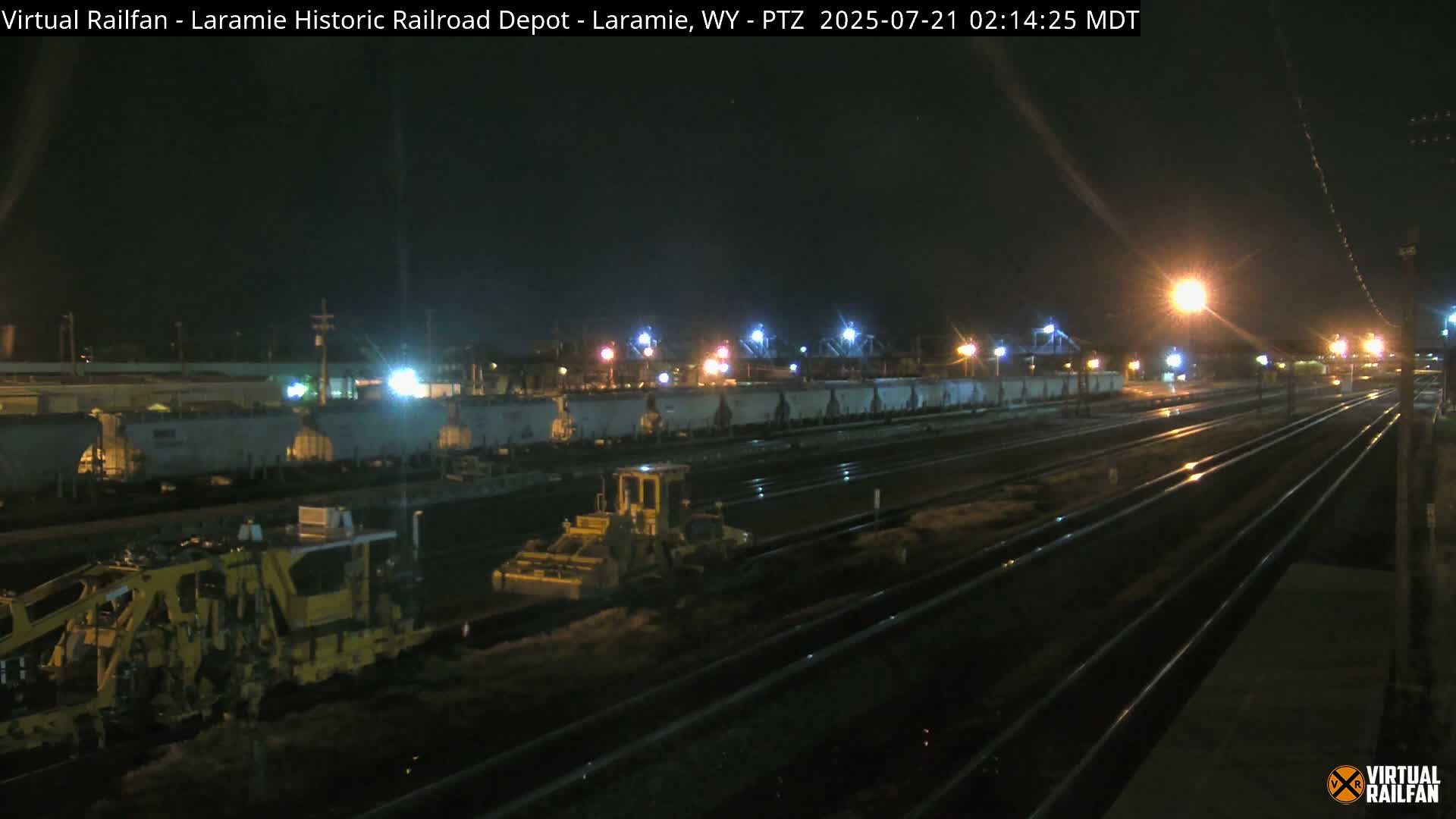 At night, under wet conditions, a yellow piece of railway maintenance equipment works near a long line of train cars parked on railroad tracks.