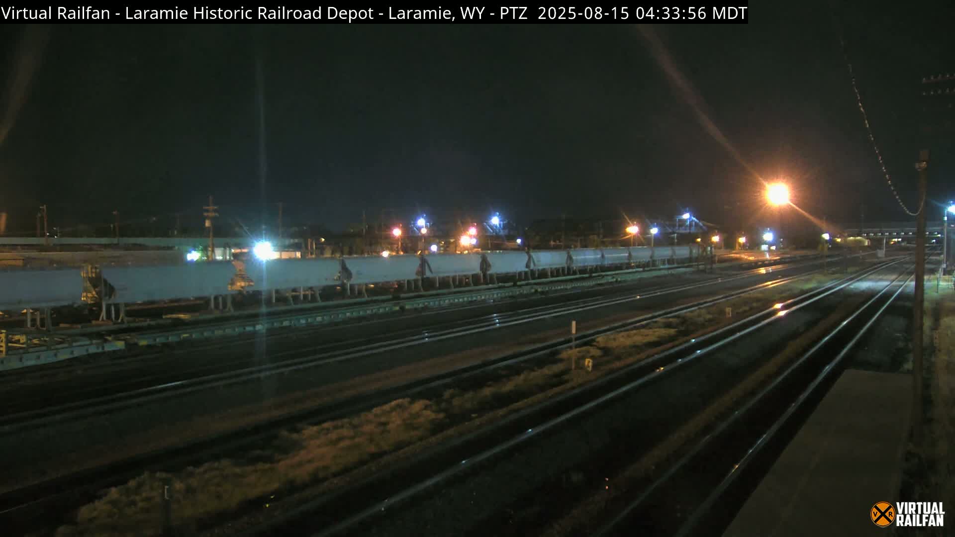 A nighttime view of railroad tracks and a depot, illuminated by streetlights, with clear skies.