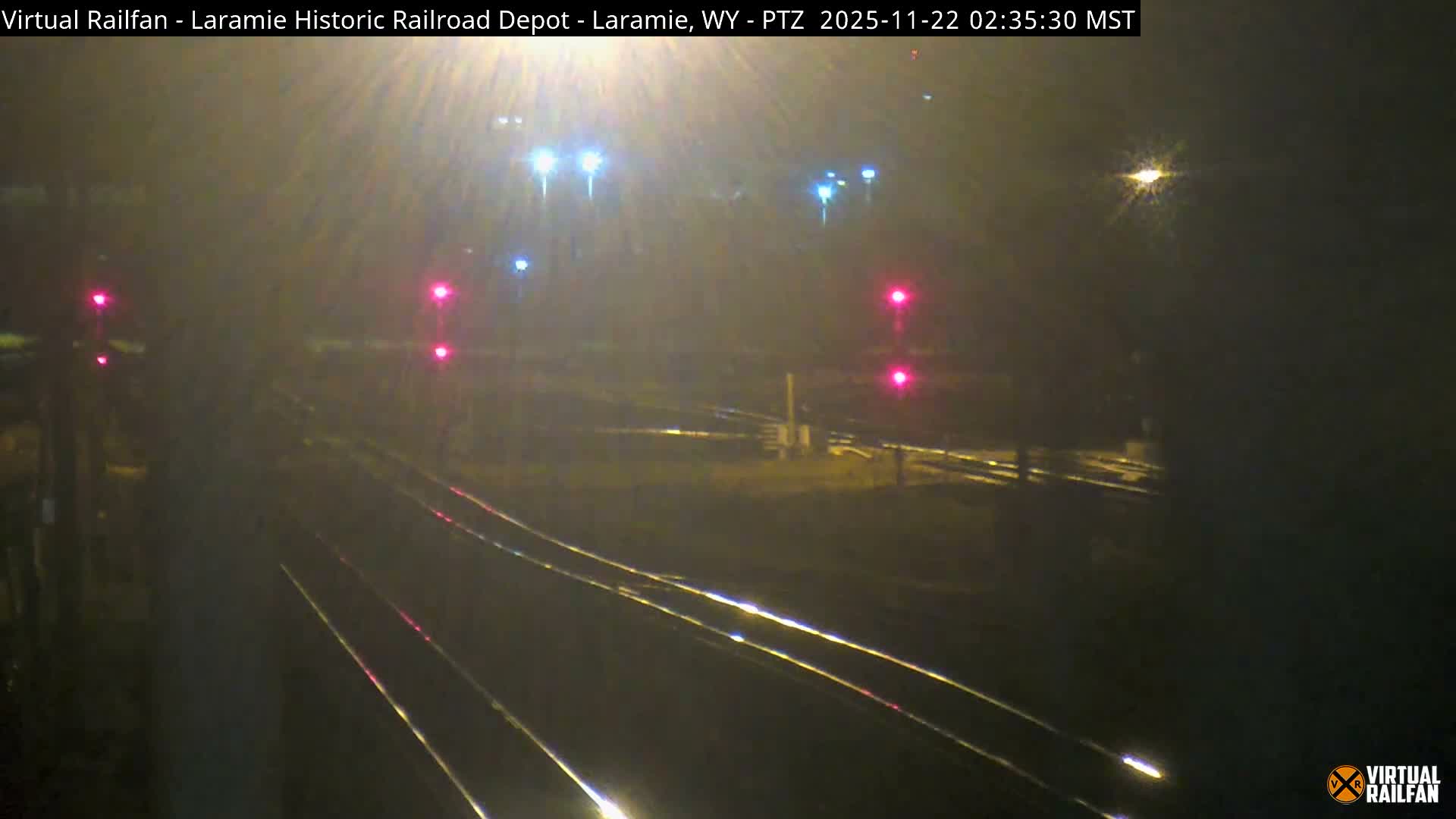 The image shows a foggy, nighttime view of multiple railroad tracks illuminated by several red signal lights and diffused distant streetlights.